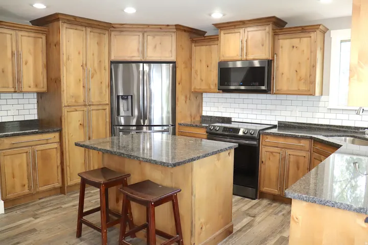 Chef's kitchen featuring custom wood cabinetry, stainless steel French-door refrigerator, black electric range, gray quartz countertops, subway tile backsplash, and a centered island with seating for two