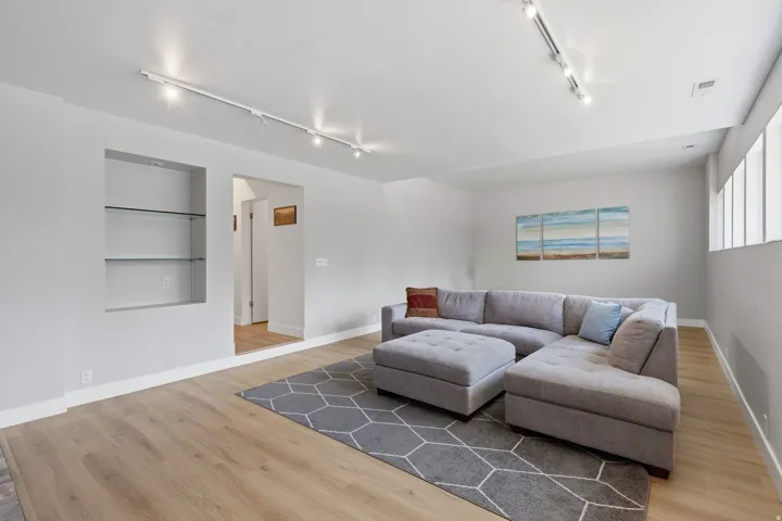 Bright, open living area featuring warm wood-finish flooring, recessed and track lighting throughout, and expansive windows providing natural light