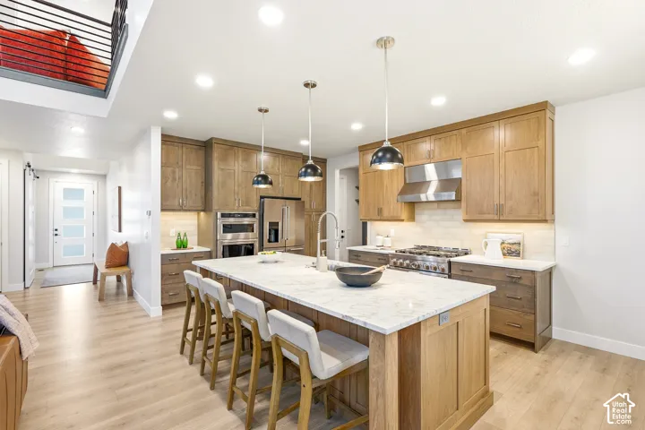 Open-concept kitchen featuring light wood-finish flooring, a large central island with a light-toned countertop, pendant lighting, stainless steel appliances, and wood cabinetry