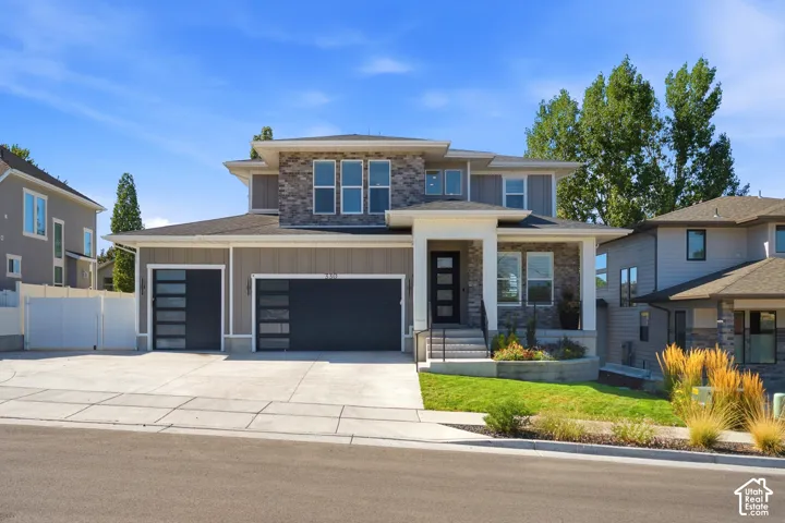 Contemporary exterior design featuring stone and siding accents, a covered entry porch, multiple garage doors with frosted glass panels, an extended concrete driveway, and mature landscaping