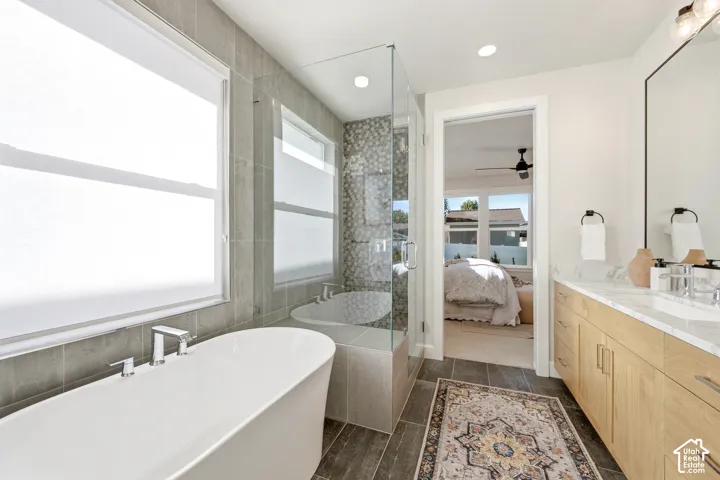 Freestanding soaking tub, glass-enclosed shower with pebble tile, dual vanity with light-tone wood cabinetry, white countertop, and dark wood-look tile flooring