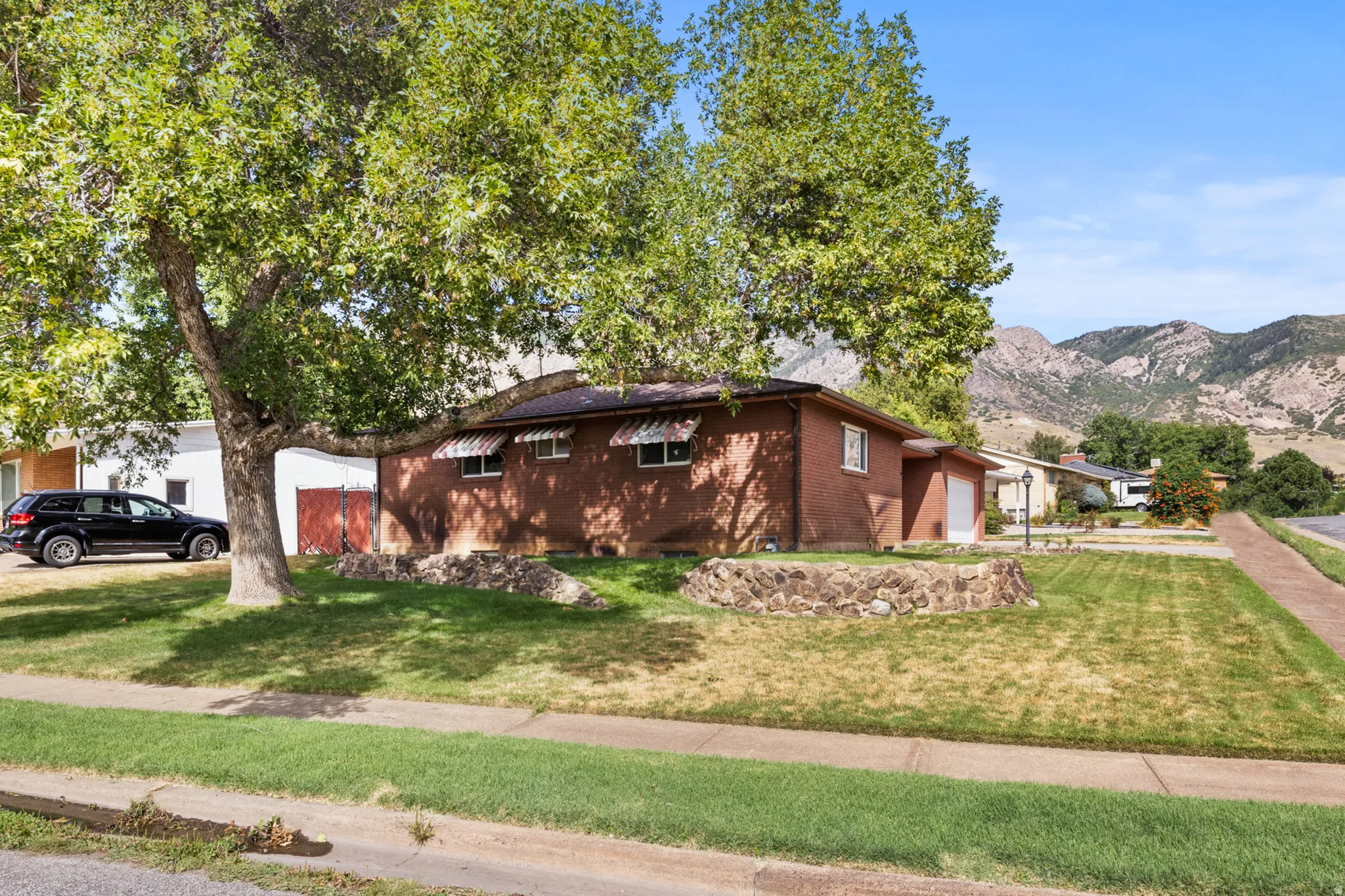Brick residence situated on a spacious lawn with mature shade trees, mountain backdrop, and single-car garage