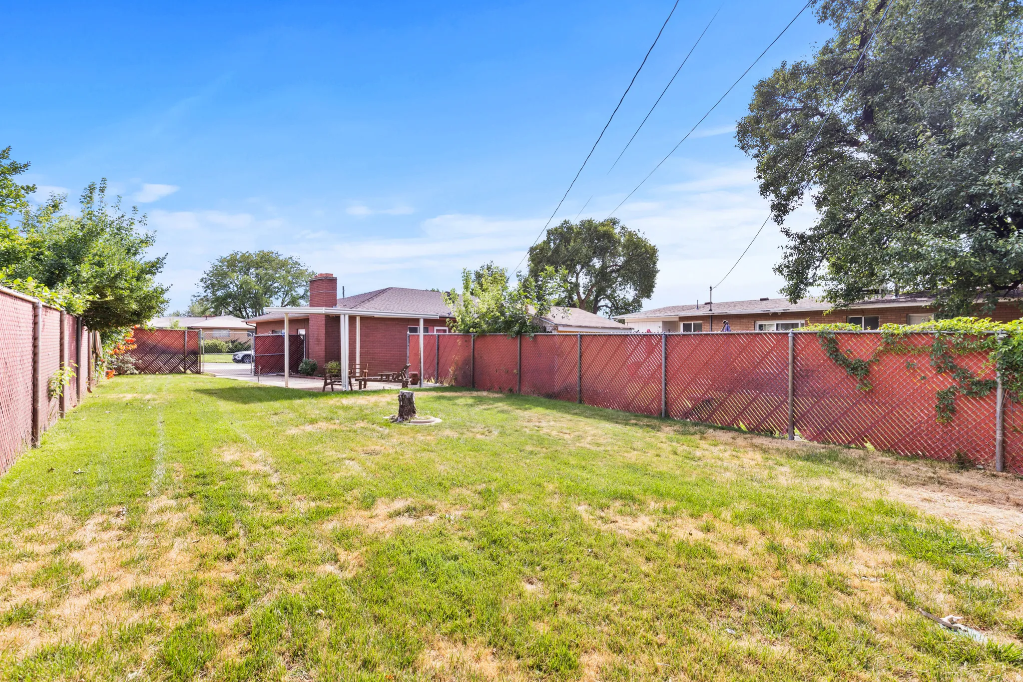 Spacious backyard featuring a lush, manicured lawn with mature shade trees, red metal privacy fencing, and detached outbuildings including a storage shed