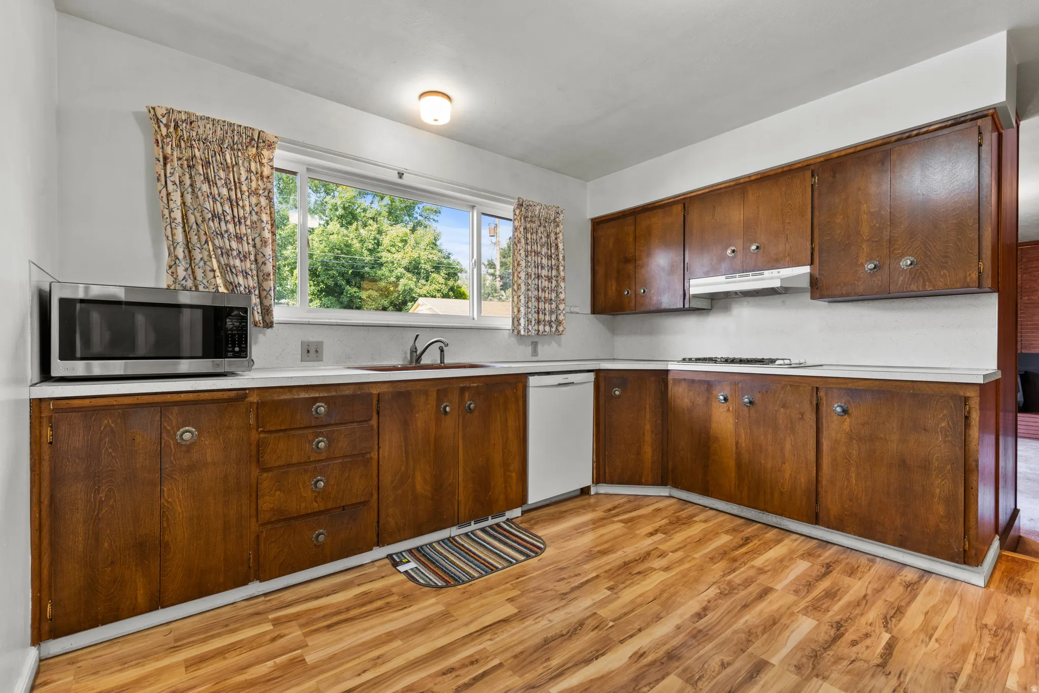 Spacious kitchen featuring rich wood cabinetry with ample storage, wood-finish flooring throughout, white countertops, stainless steel appliances, and abundant natural light from a large window with custom drapery