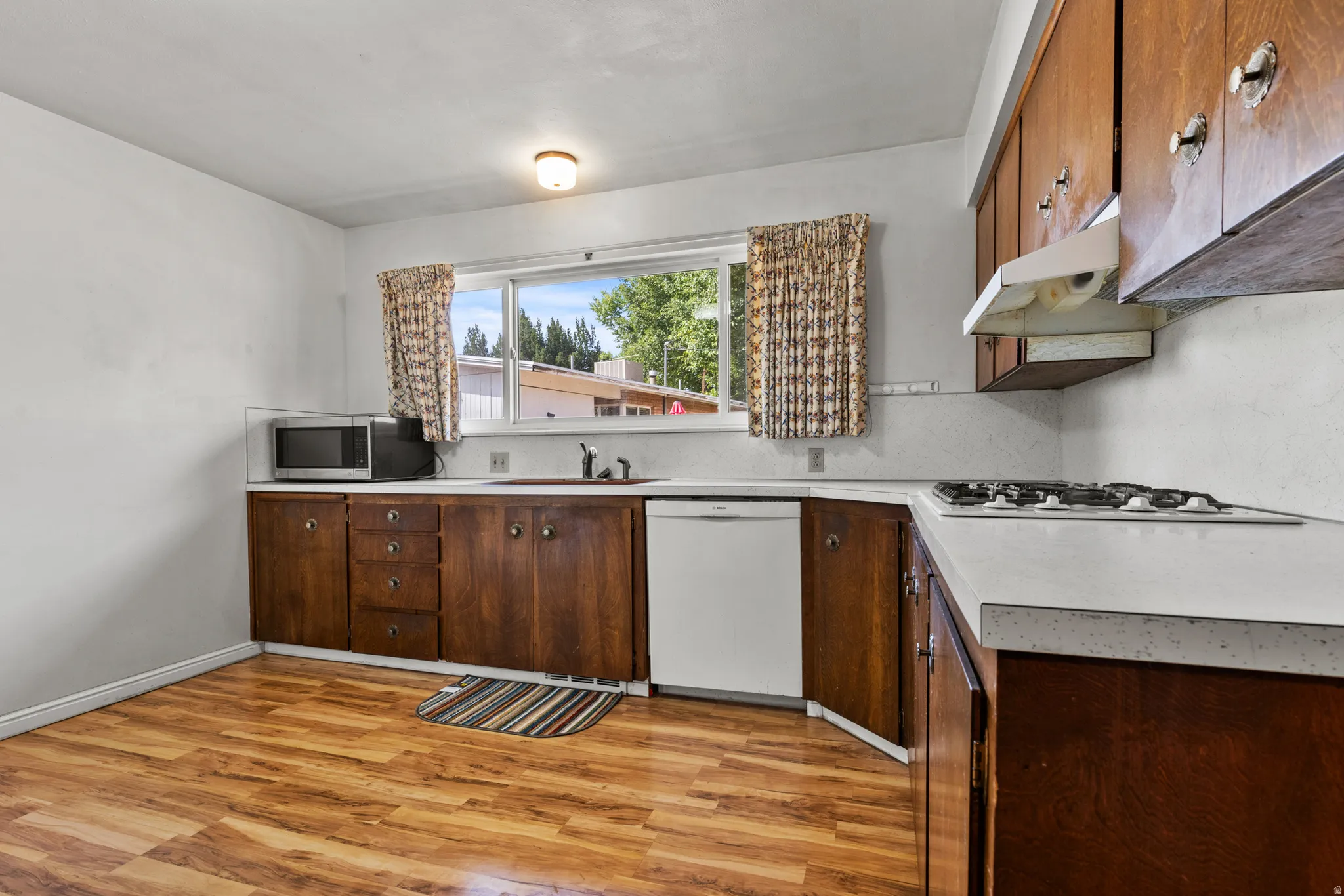 Galley kitchen featuring warm wood-finish cabinetry, stainless steel appliances including a gas cooktop and dishwasher, wood-finish flooring, and a window overlooking natural scenery