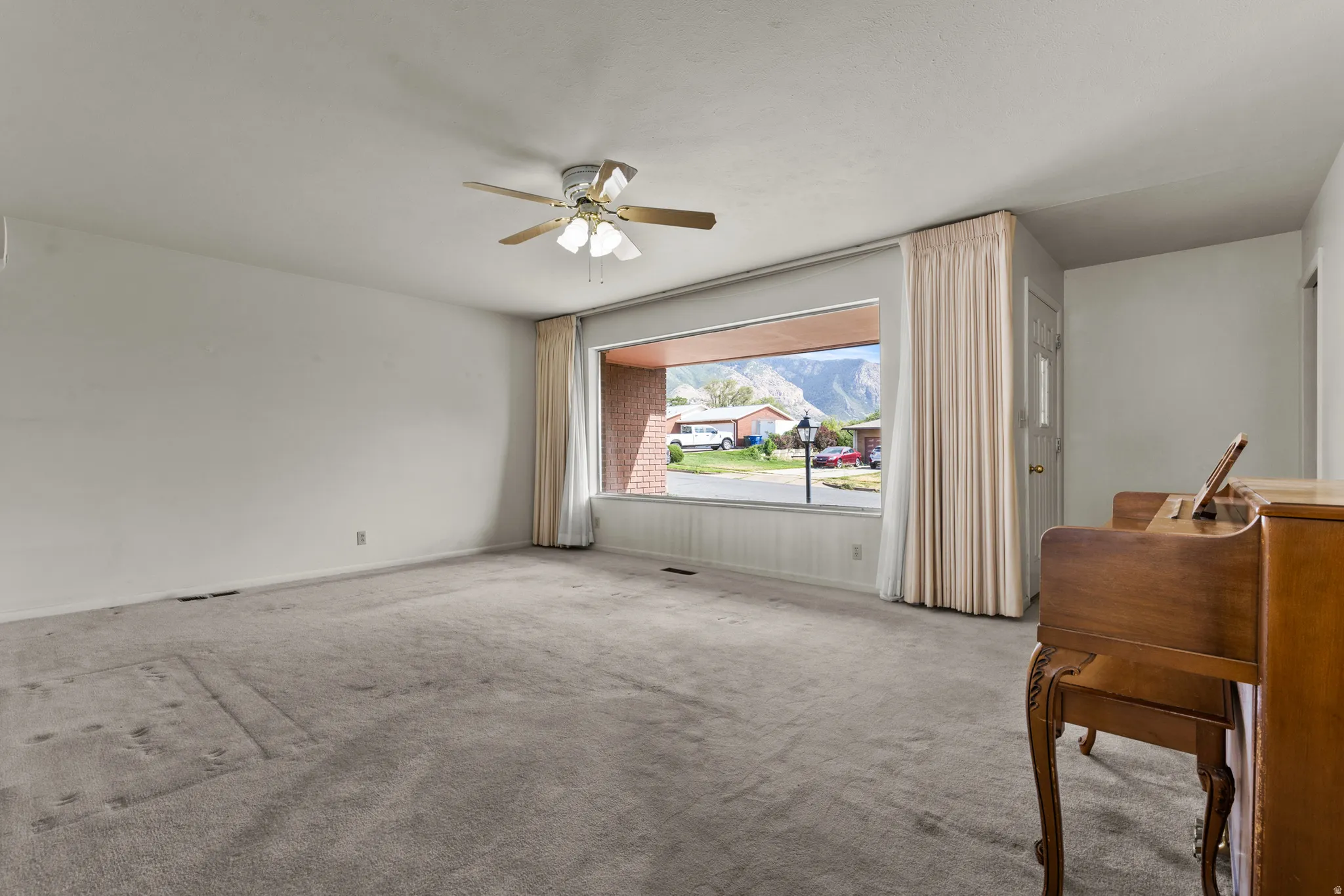 Spacious room featuring neutral carpeting, large picture window with natural light, ceiling fan with integrated lighting, and wood-finish furnishings