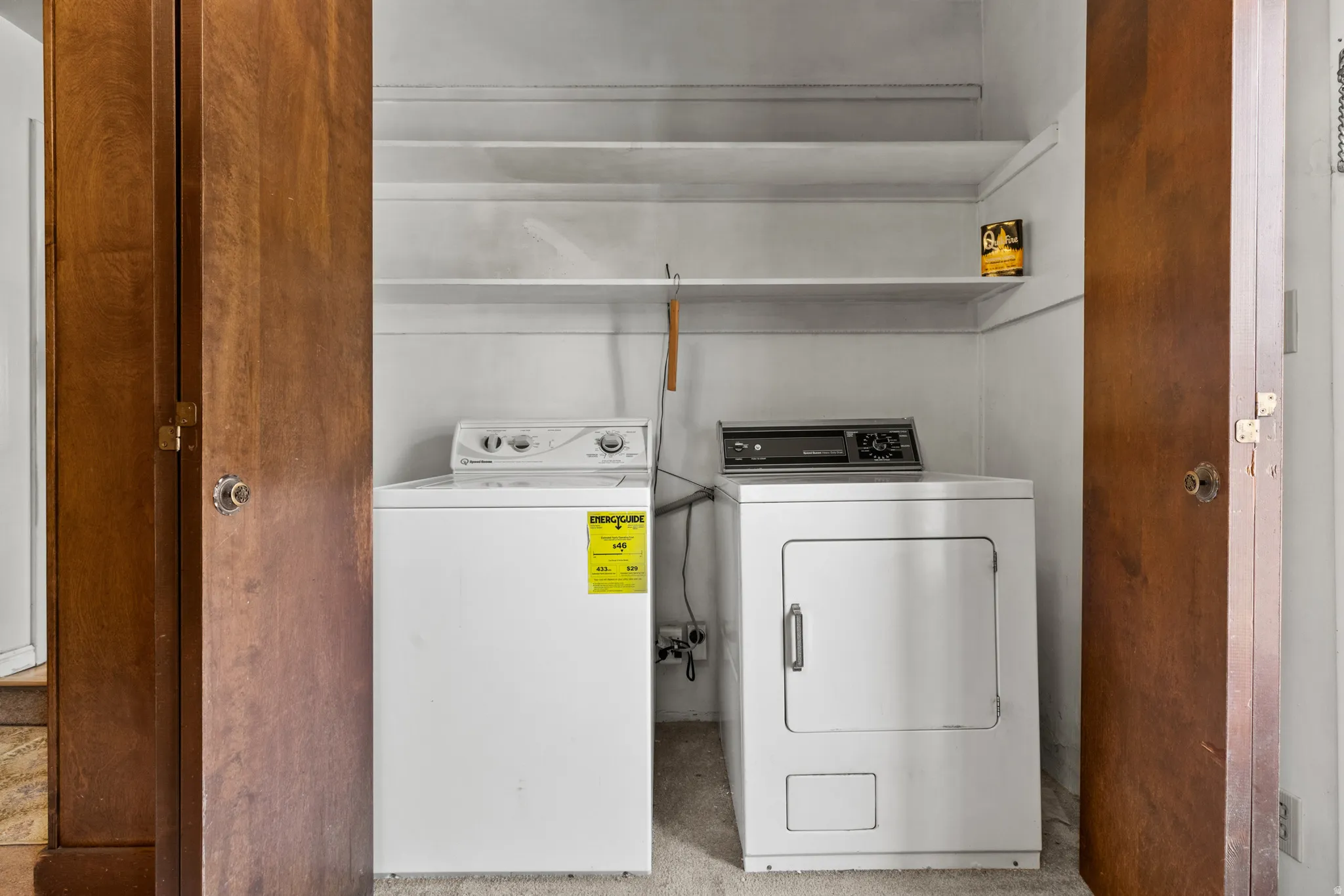 Dedicated laundry room featuring stacked washer and electric dryer with overhead shelving for storage, enclosed by wood-finish double doors for privacy and convenience