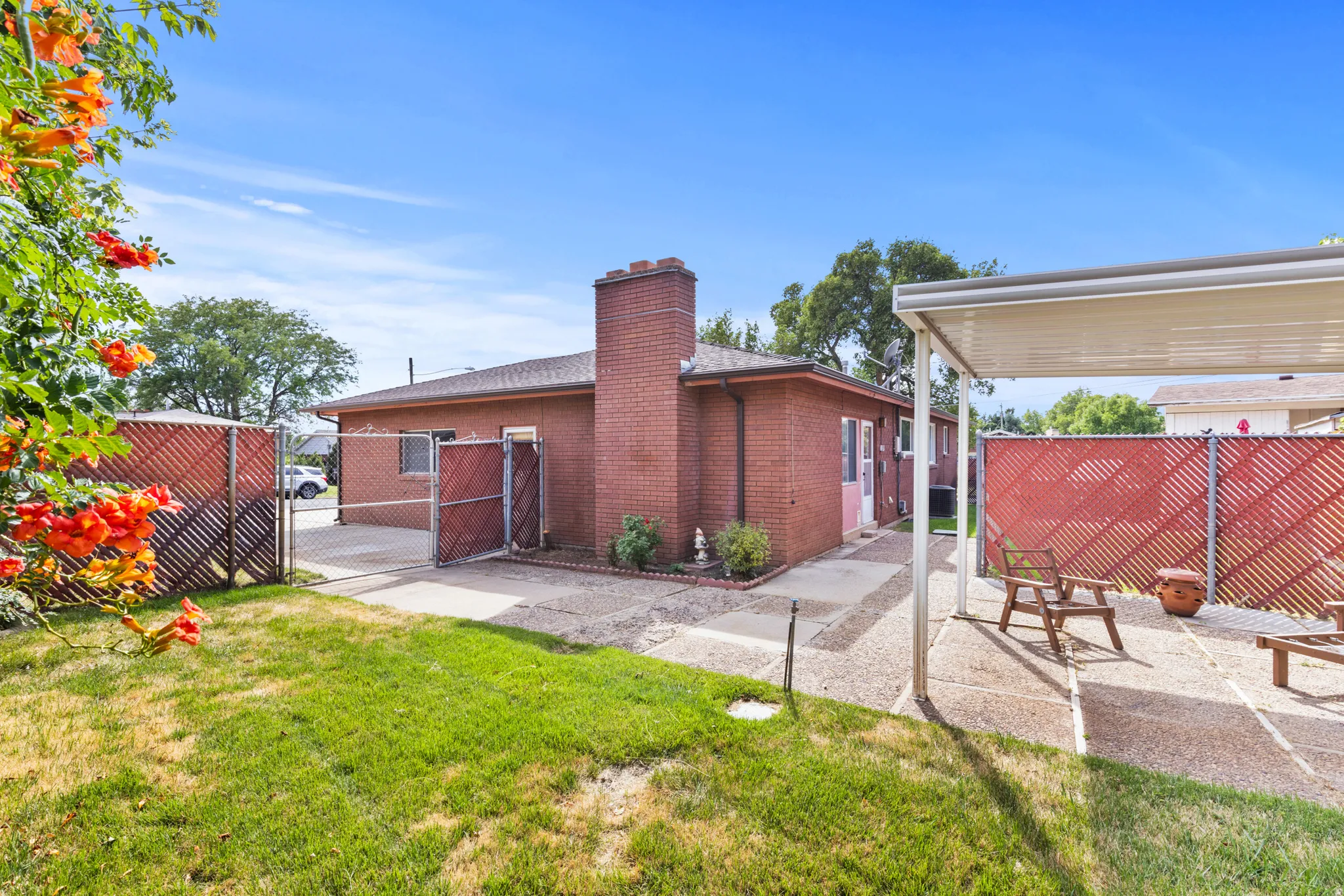 Solid brick residence with a functional covered patio featuring metal fencing, mature landscaping with flowering shrubs, and a level lawn ideal for outdoor use