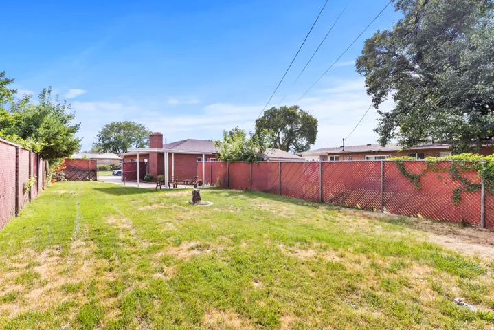 Spacious backyard featuring a lush, manicured lawn with mature shade trees, red metal privacy fencing, and detached outbuildings including a storage shed