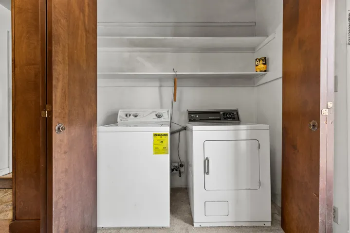 Dedicated laundry room featuring stacked washer and electric dryer with overhead shelving for storage, enclosed by wood-finish double doors for privacy and convenience