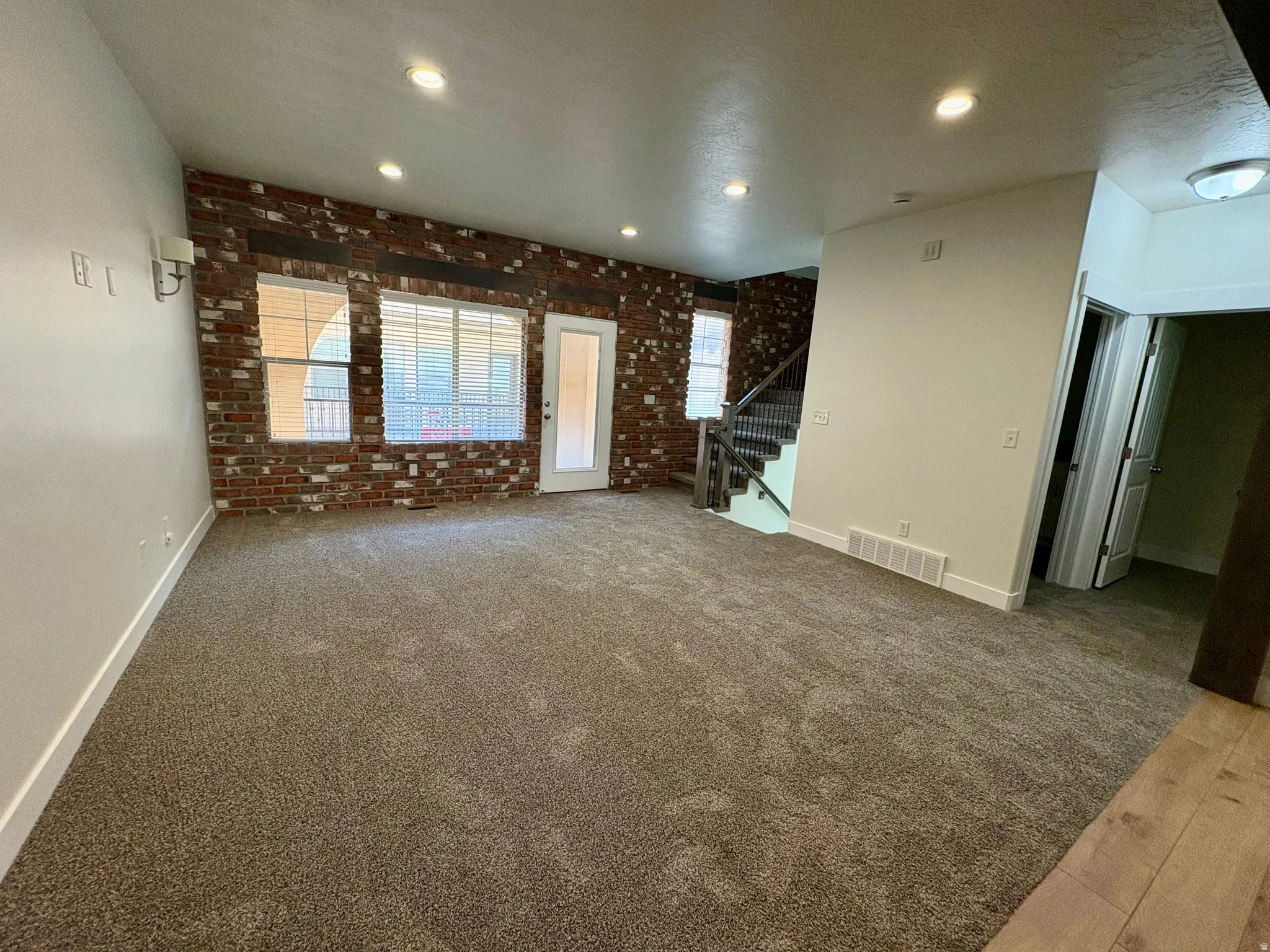 Spacious living area featuring neutral carpet flooring, exposed brick accent wall with multiple windows, recessed ceiling lighting, and glass door entry