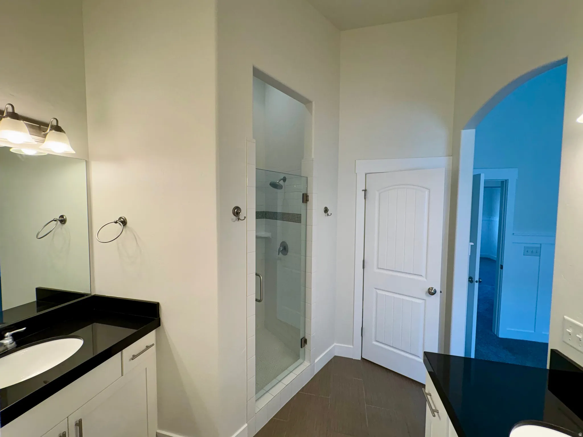 Dual vanities with dark countertops, a walk-in shower with glass enclosure, white paneled doors, and arched doorways