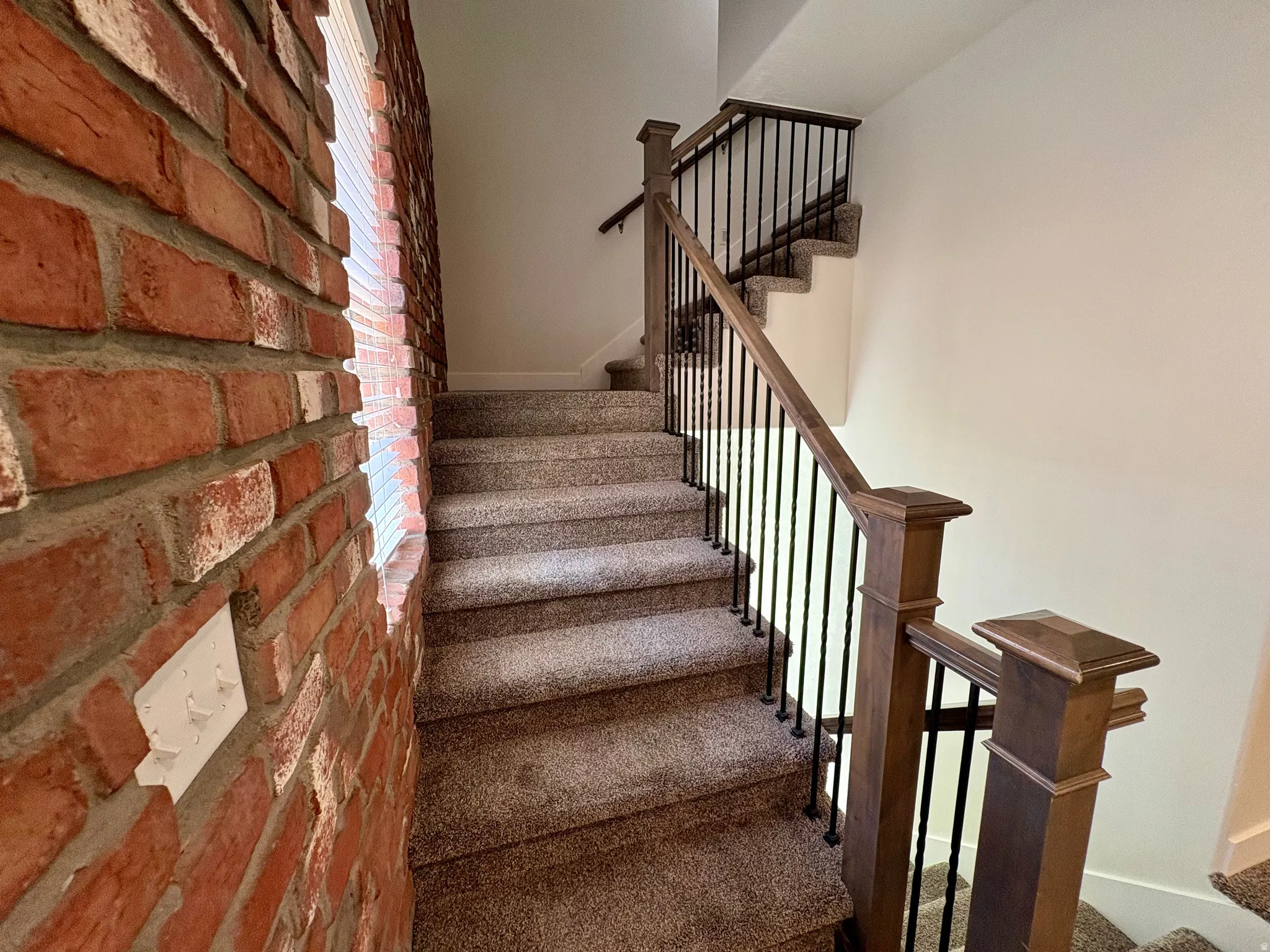 Carpeted staircase featuring a wood railing with black metal balusters and a brick accent wall