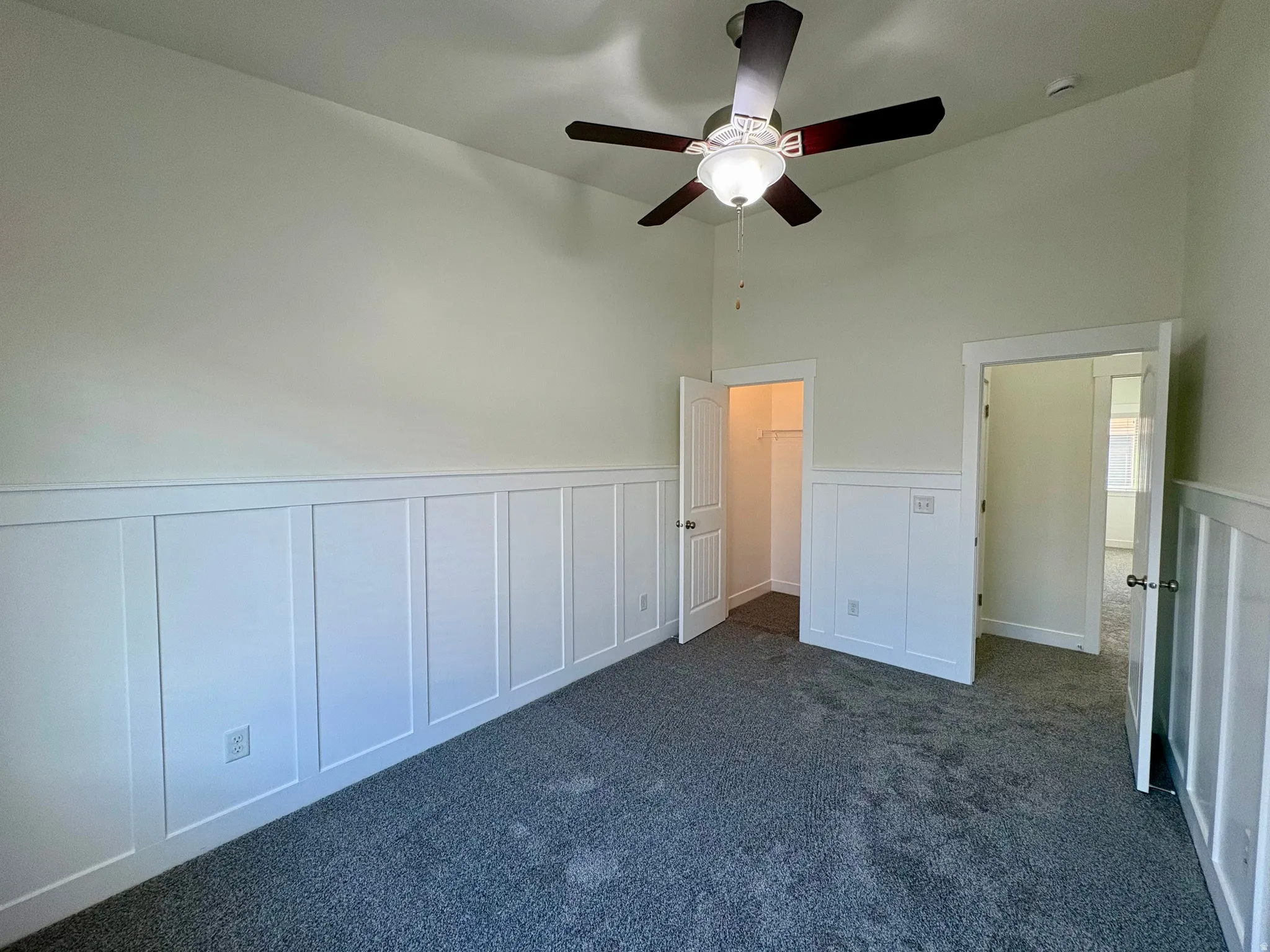 Spacious room featuring wainscoting detail along lower walls, neutral paint finishes, carpet flooring, and ceiling fan with integrated lighting