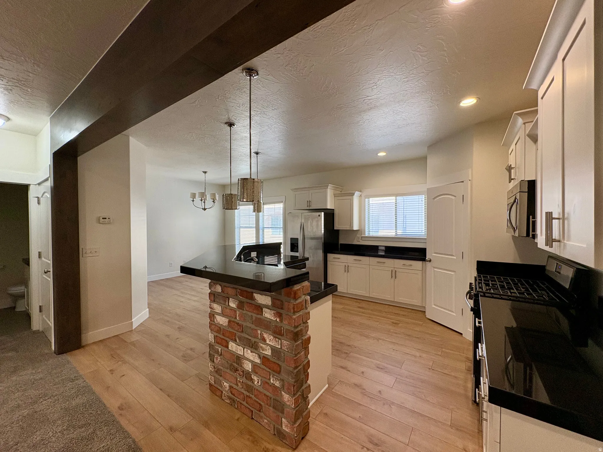 Kitchen featuring white cabinetry, black countertops, stainless steel appliances, wood-finish flooring, and a brick-clad island with pendant lighting