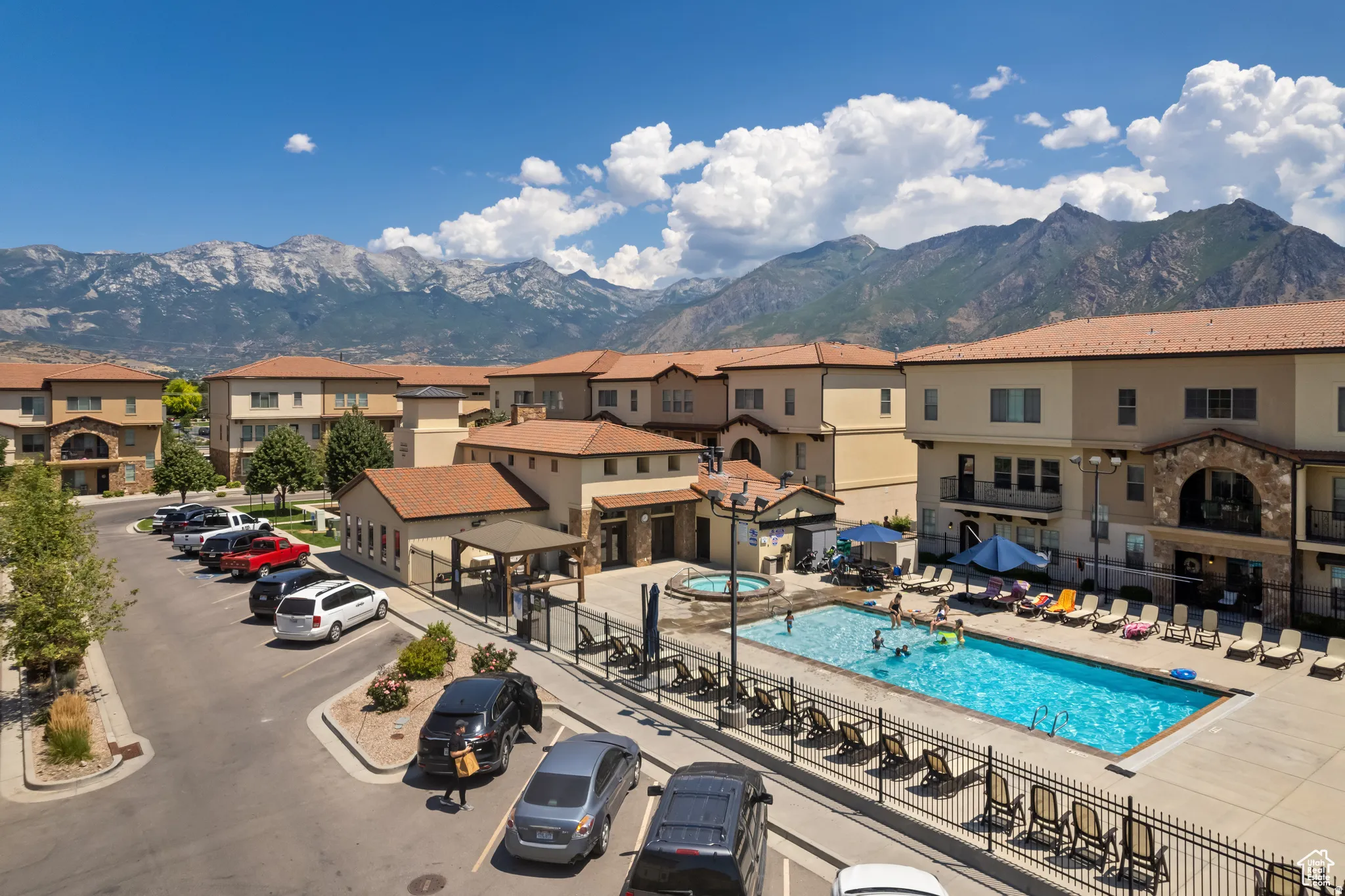 Resort-style community pool with lounge seating and fencing, complemented by Mediterranean-inspired architecture featuring terracotta tile roofing, stucco exterior finishes, and multi-unit residential buildings set against mountain backdrop