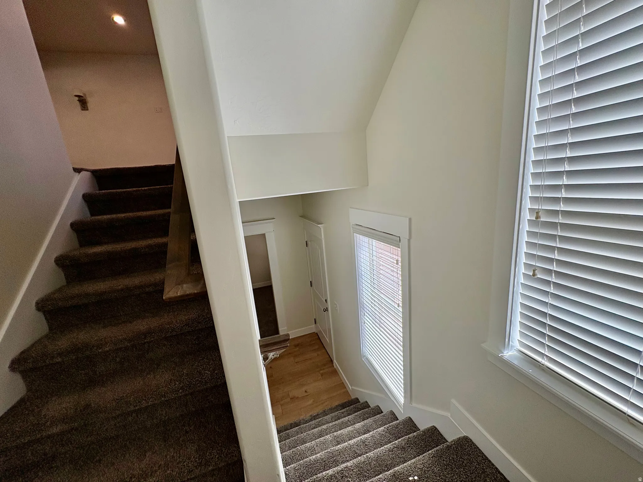 Carpeted staircase with white balustrade, featuring a window with horizontal blinds and wood-finish flooring visible below
