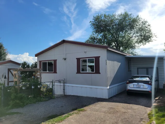 Light-colored siding exterior with contrasting trim windows, covered carport, gravel driveway, and a chain-link fence