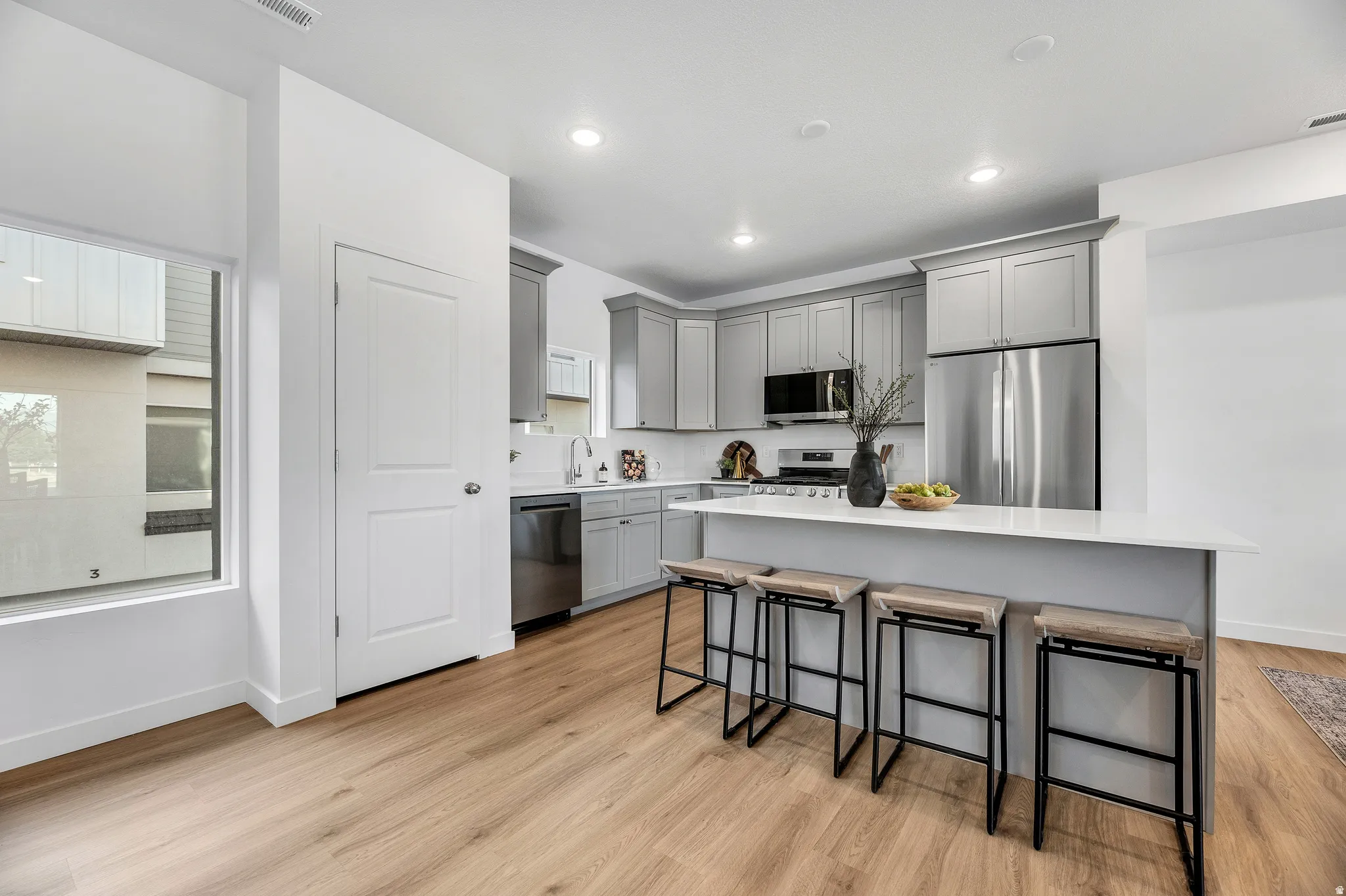 Modern kitchen featuring light wood-finish flooring, stainless steel appliances, grey cabinetry, a white countertop island, and recessed lighting
