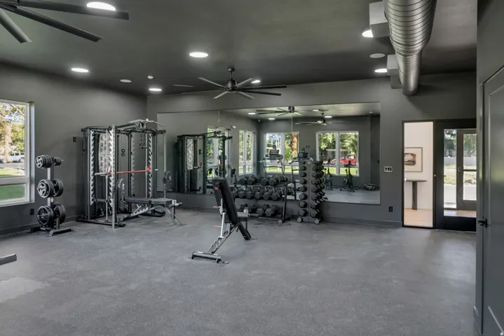 State-of-the-art fitness facility featuring multiple strength training stations with glass enclosures, polished concrete flooring, recessed and pendant ceiling lighting, ceiling fan, and expansive windows overlooking landscaped grounds