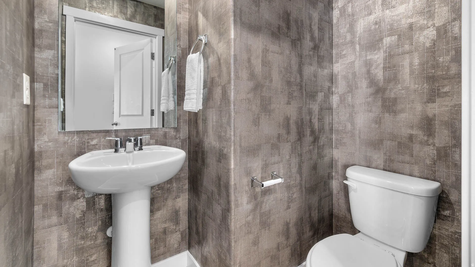 Pedestal sink with chrome fixtures, white toilet, and textured gray wall covering