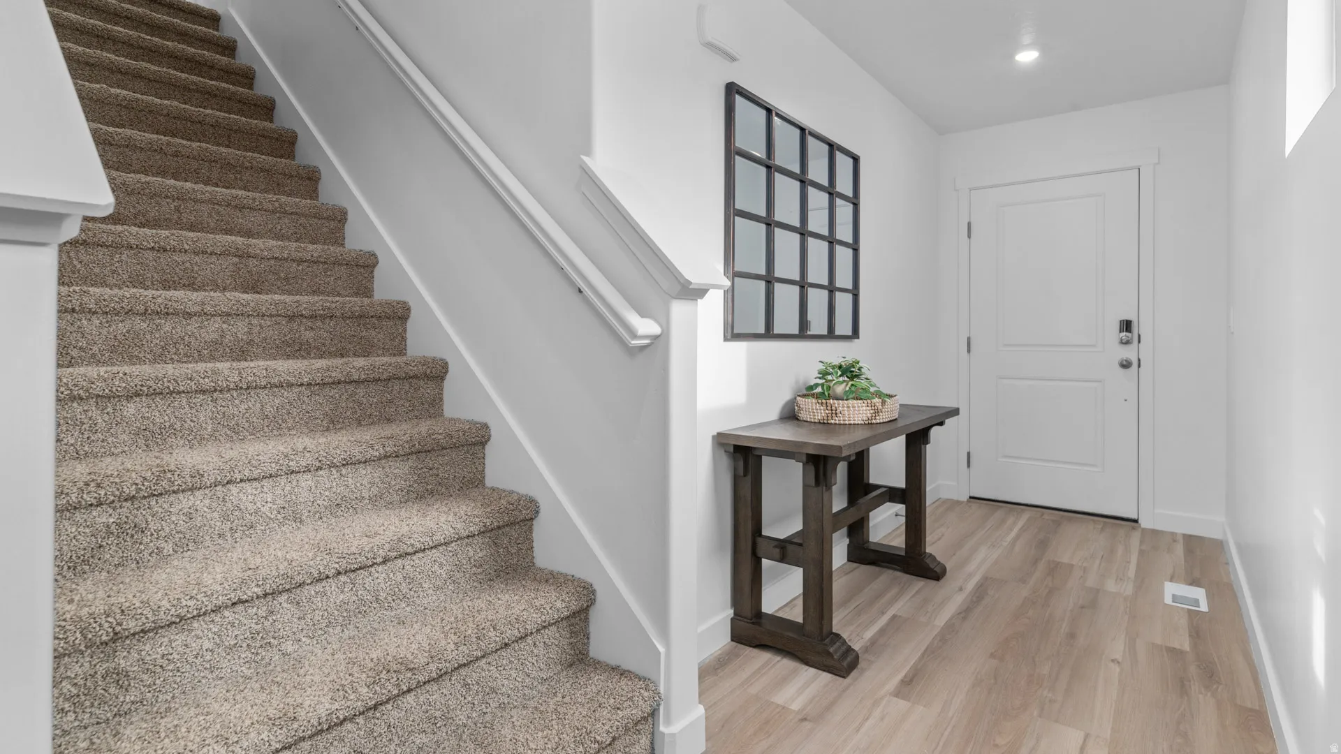 Carpeted staircase with white handrail, light wood-finish flooring, white entry door with smart lock, and recessed lighting