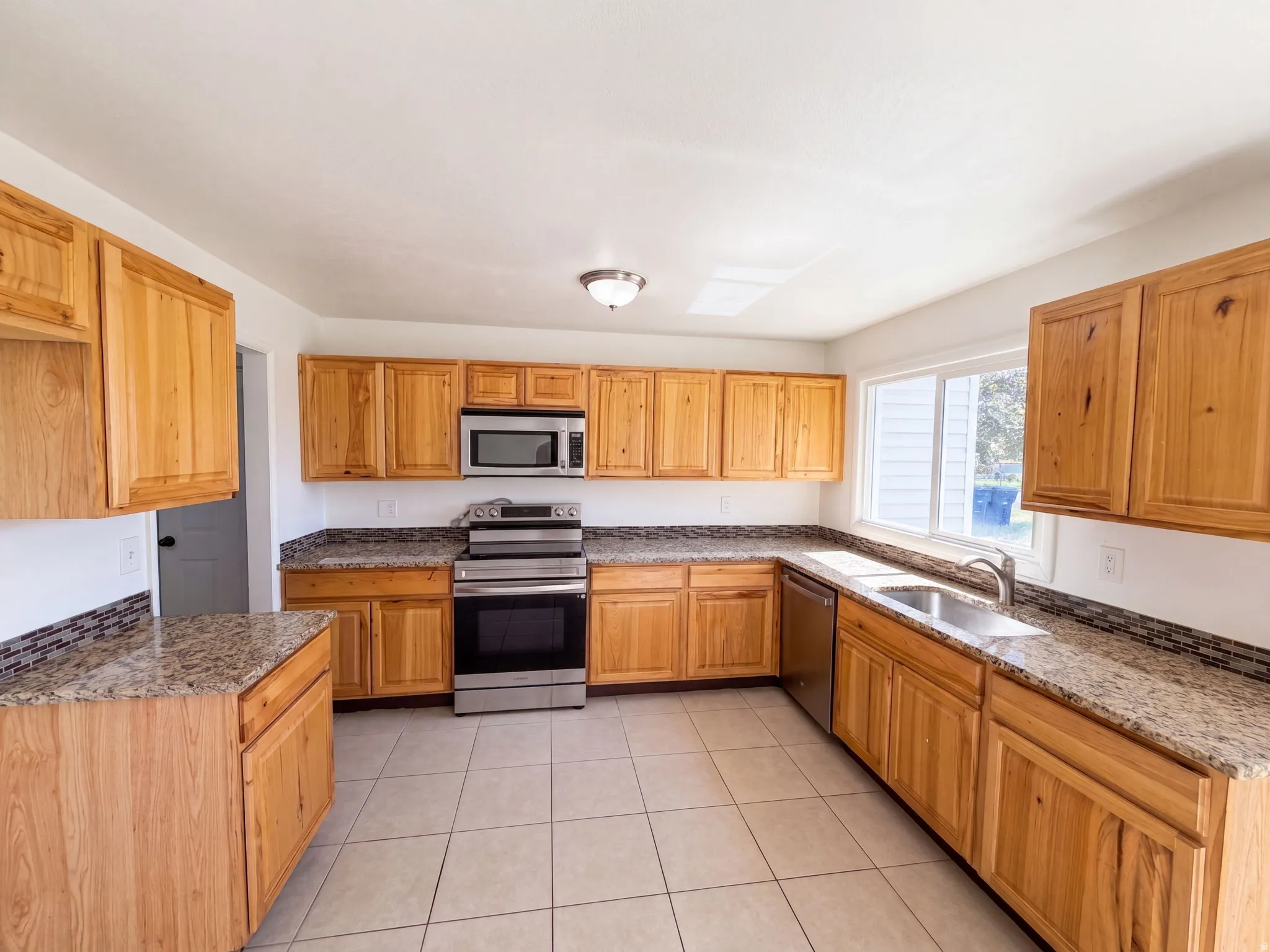 Spacious kitchen featuring wood cabinetry, granite countertops, stainless steel appliances including a gas range and microwave, ceramic tile flooring, and abundant natural light from multiple windows