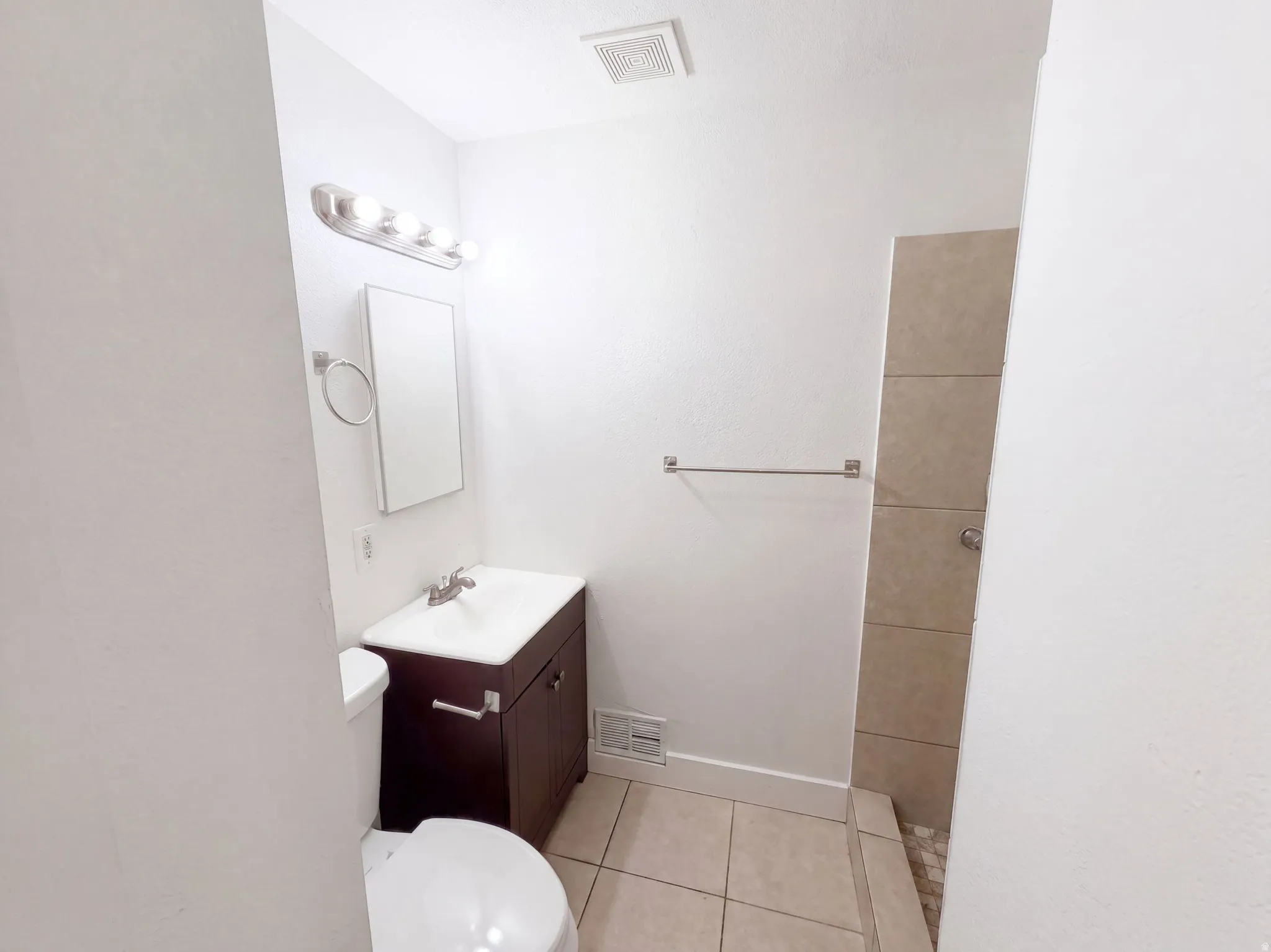 Compact bathroom featuring a dark wood-finish vanity with integrated sink, ceramic tile flooring, and a frameless glass shower enclosure with brushed nickel hardware