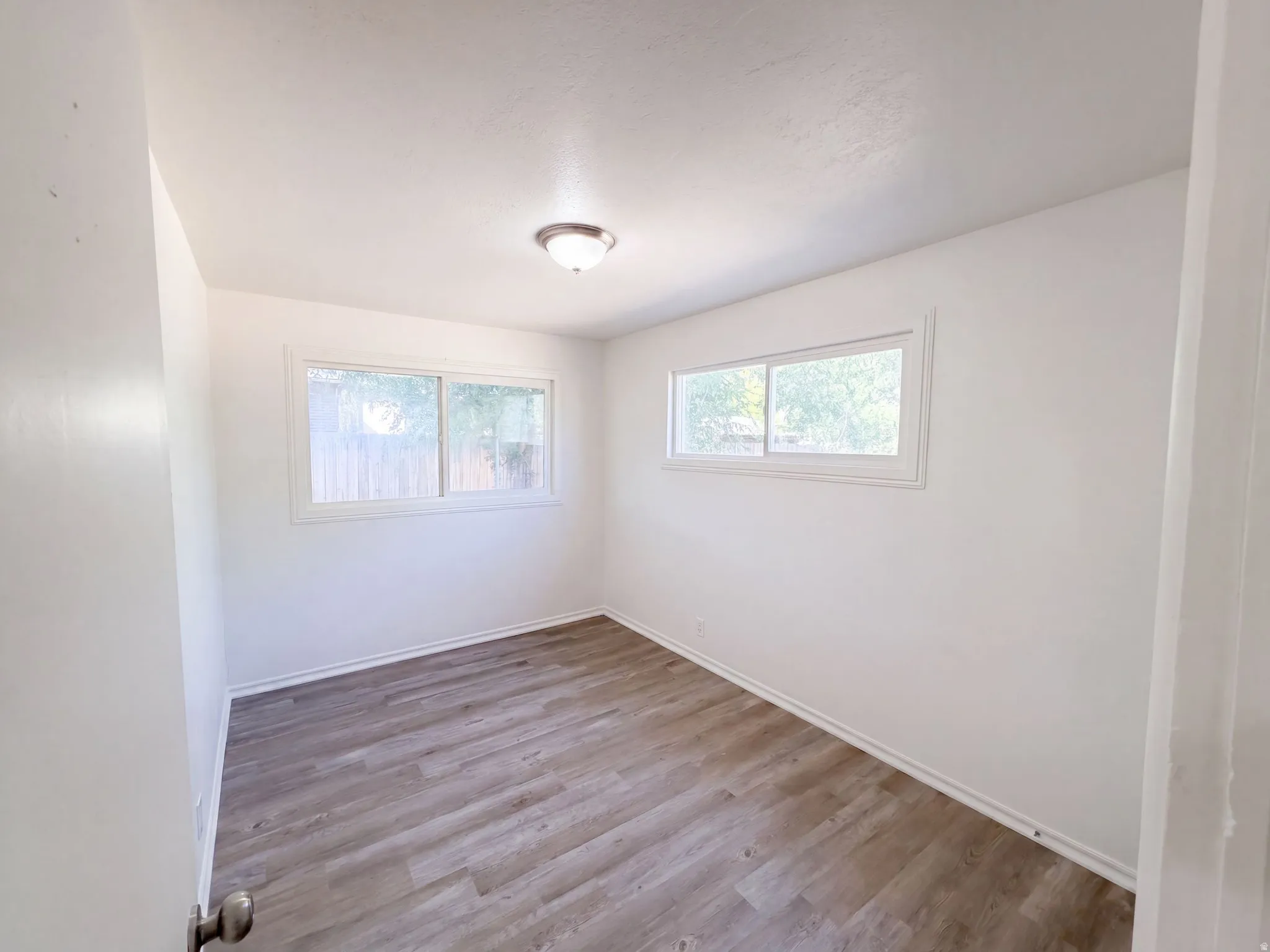 Bright, light-filled bedroom with dual windows providing natural illumination, wood-finish flooring throughout, and clean white walls with recessed ceiling lighting