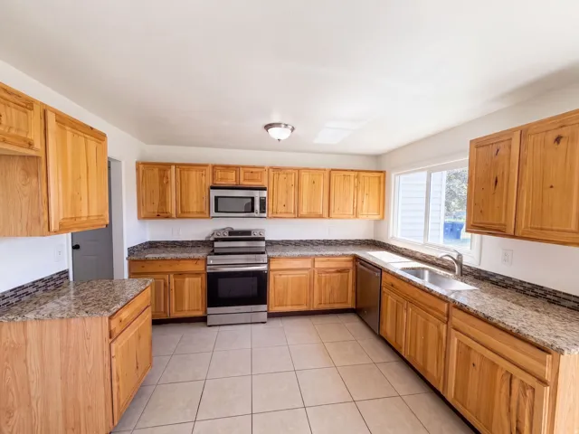 Spacious kitchen featuring wood cabinetry, granite countertops, stainless steel appliances including a gas range and microwave, ceramic tile flooring, and abundant natural light from multiple windows