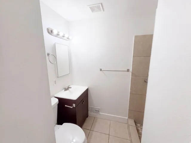 Compact bathroom featuring a dark wood-finish vanity with integrated sink, ceramic tile flooring, and a frameless glass shower enclosure with brushed nickel hardware