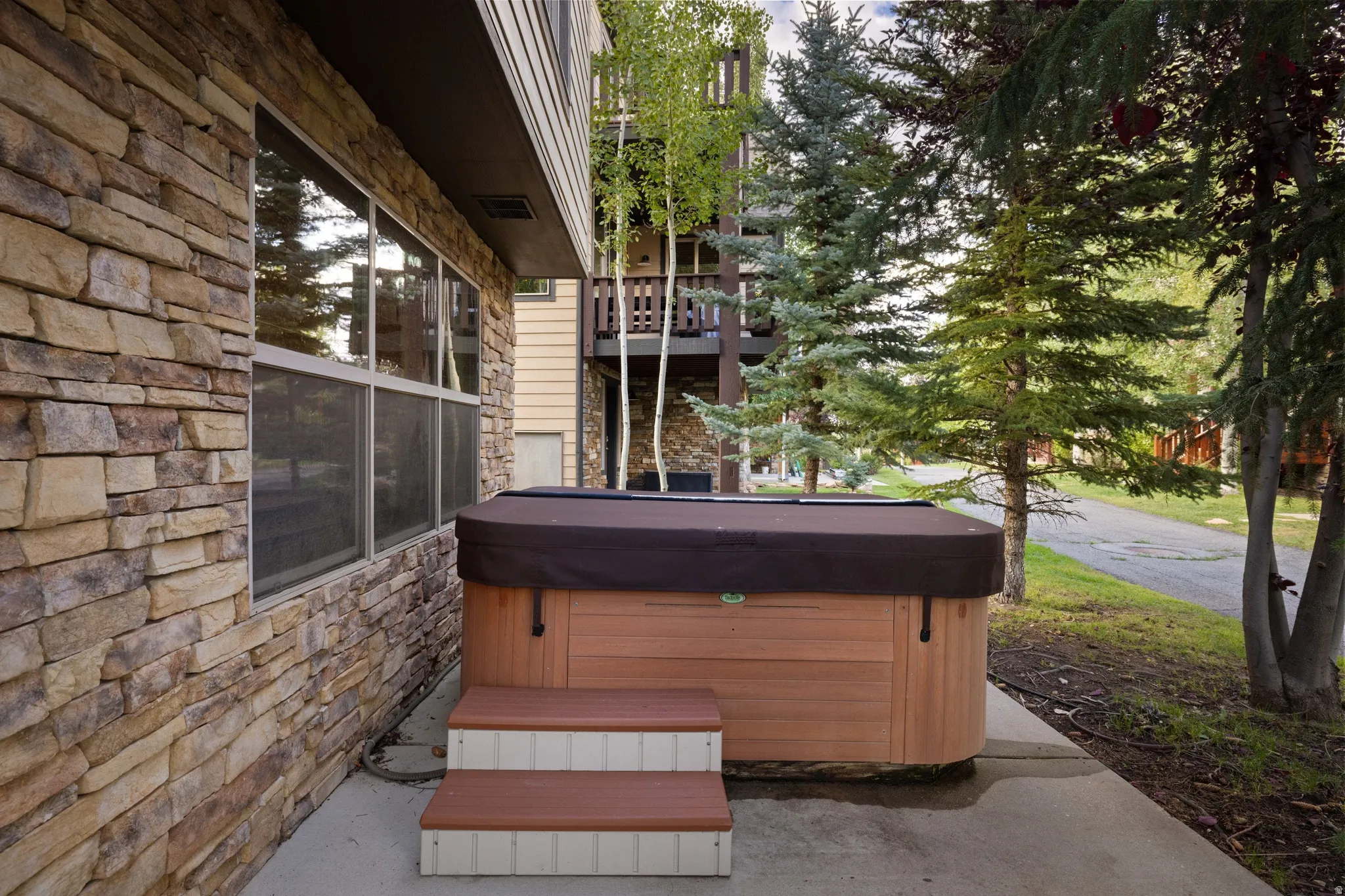 Spa-inspired hot tub with dark cover and cushioned seating on a concrete patio, framed by natural stone siding and mature evergreen landscaping