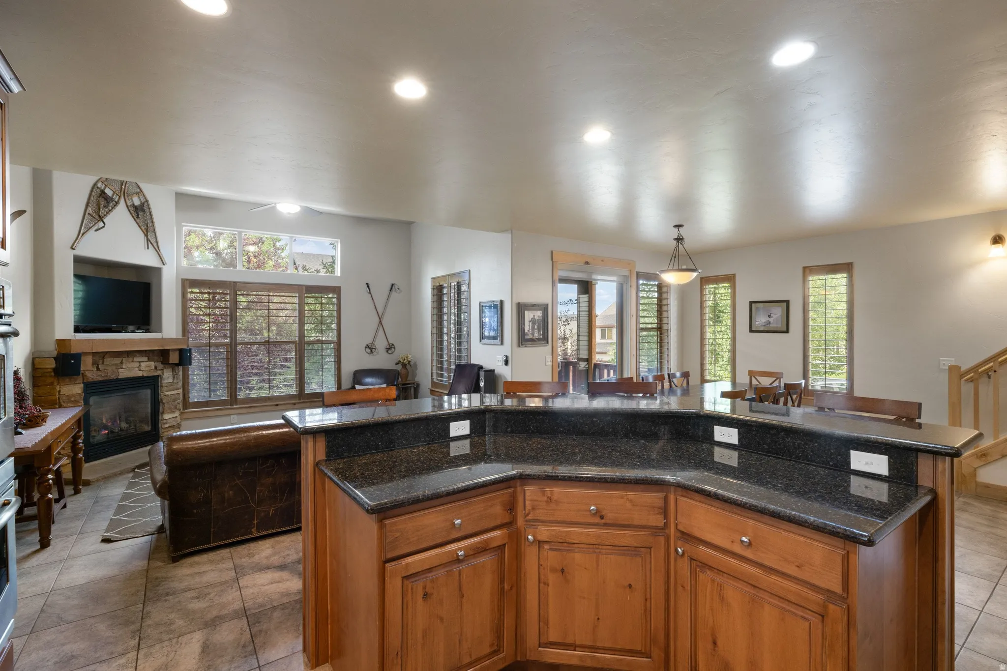 Open-concept kitchen and living area featuring warm wood cabinetry with dark stone countertops, recessed ceiling lighting, tile flooring, a fireplace with stone surround, and multiple windows with plantation shutters providing natural illumination through