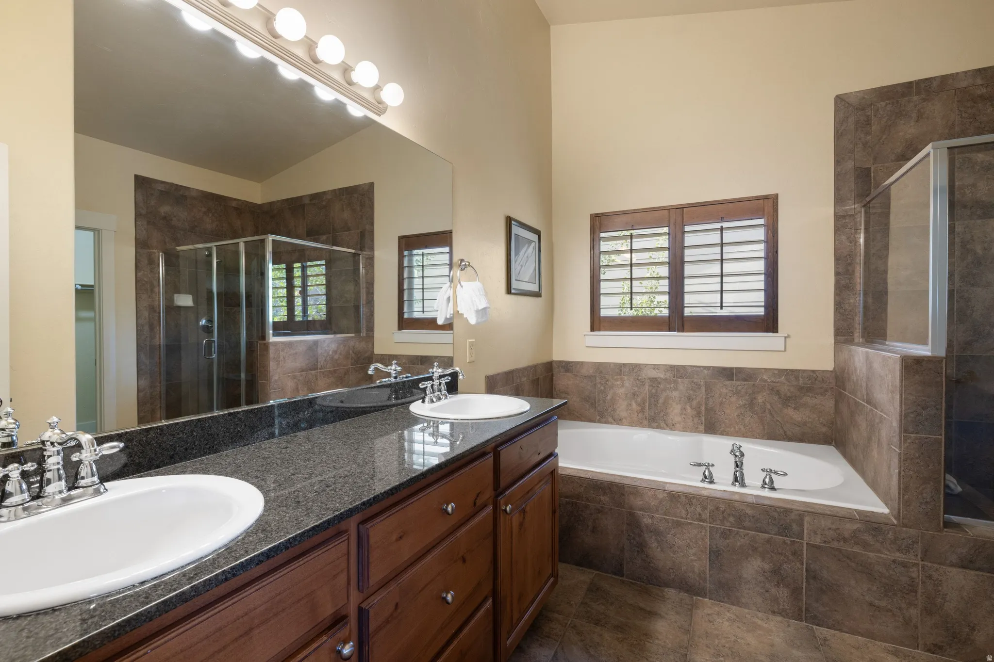 Spa-inspired master bathroom featuring dual vanities with wood cabinetry and dark stone countertops, a soaking tub with tile surround, frameless glass shower enclosure, and chrome fixtures throughout