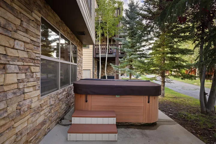 Spa-inspired hot tub with dark cover and cushioned seating on a concrete patio, framed by natural stone siding and mature evergreen landscaping