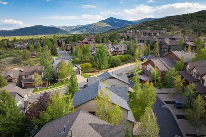 Aerial view showcasing a multi-home residential community nestled against mountain backdrop, featuring a mix of contemporary architecture with varied rooflines, asphalt driveways, mature landscaping with native trees throughout the development, and manicu