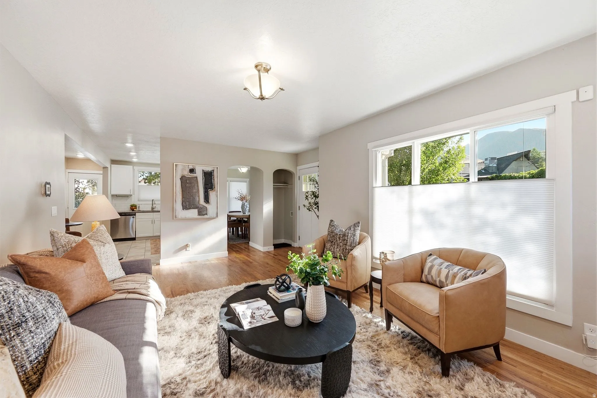 Bright, light-filled living room showcasing hardwood flooring, large multi-pane windows with natural light, a black round accent table, a tan leather armchair, and an open floor plan that extends toward the kitchen and dining areas
