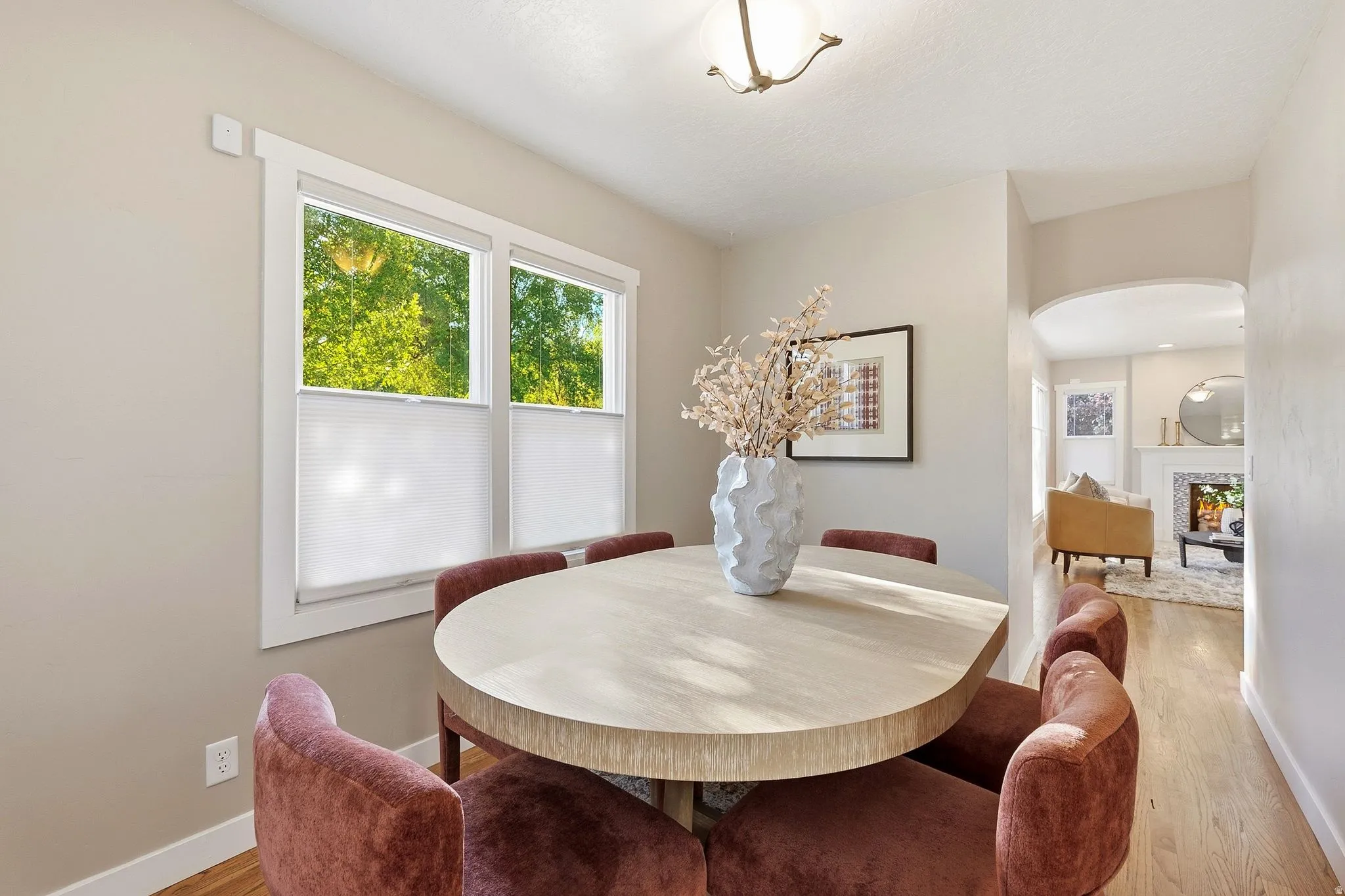 Elegant dining space featuring a light wood-finish round table, warm terracotta upholstered seating, and abundant natural light from dual casement windows