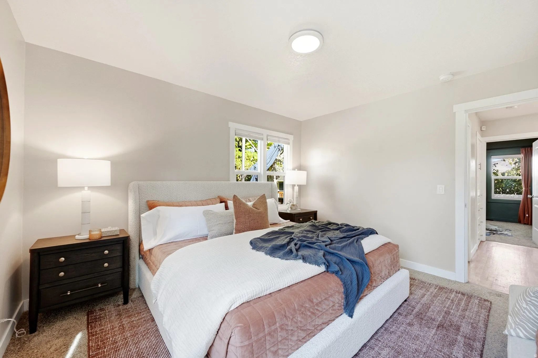 Bright secondary bedroom featuring recessed ceiling lighting, dual bedside table lamps with integrated nightstands, large windows with natural light, neutral wall finishes, and patterned area rugging