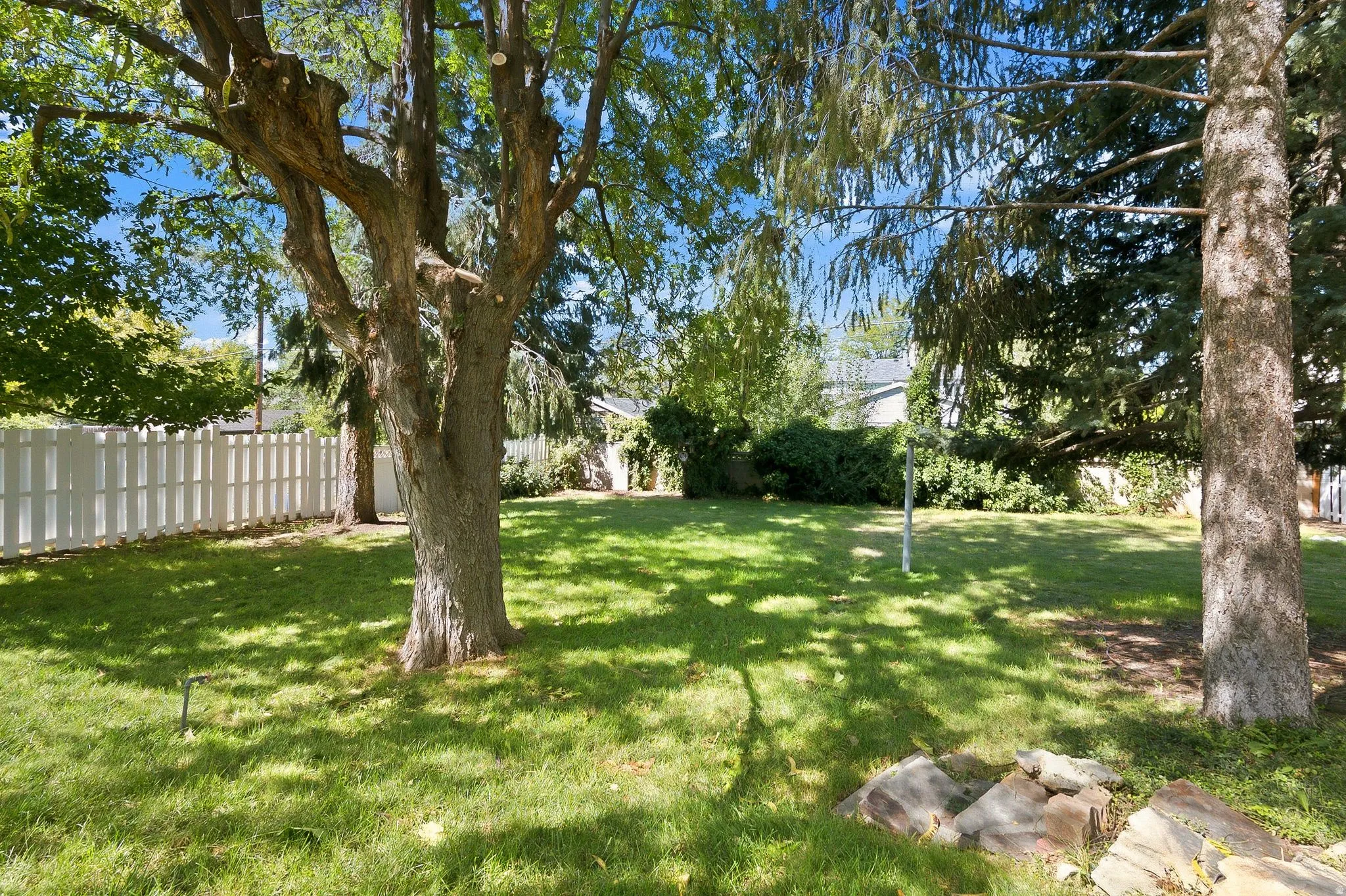 Expansive backyard featuring mature shade trees, lush manicured lawn, white picket fencing, and natural stone landscaping elements