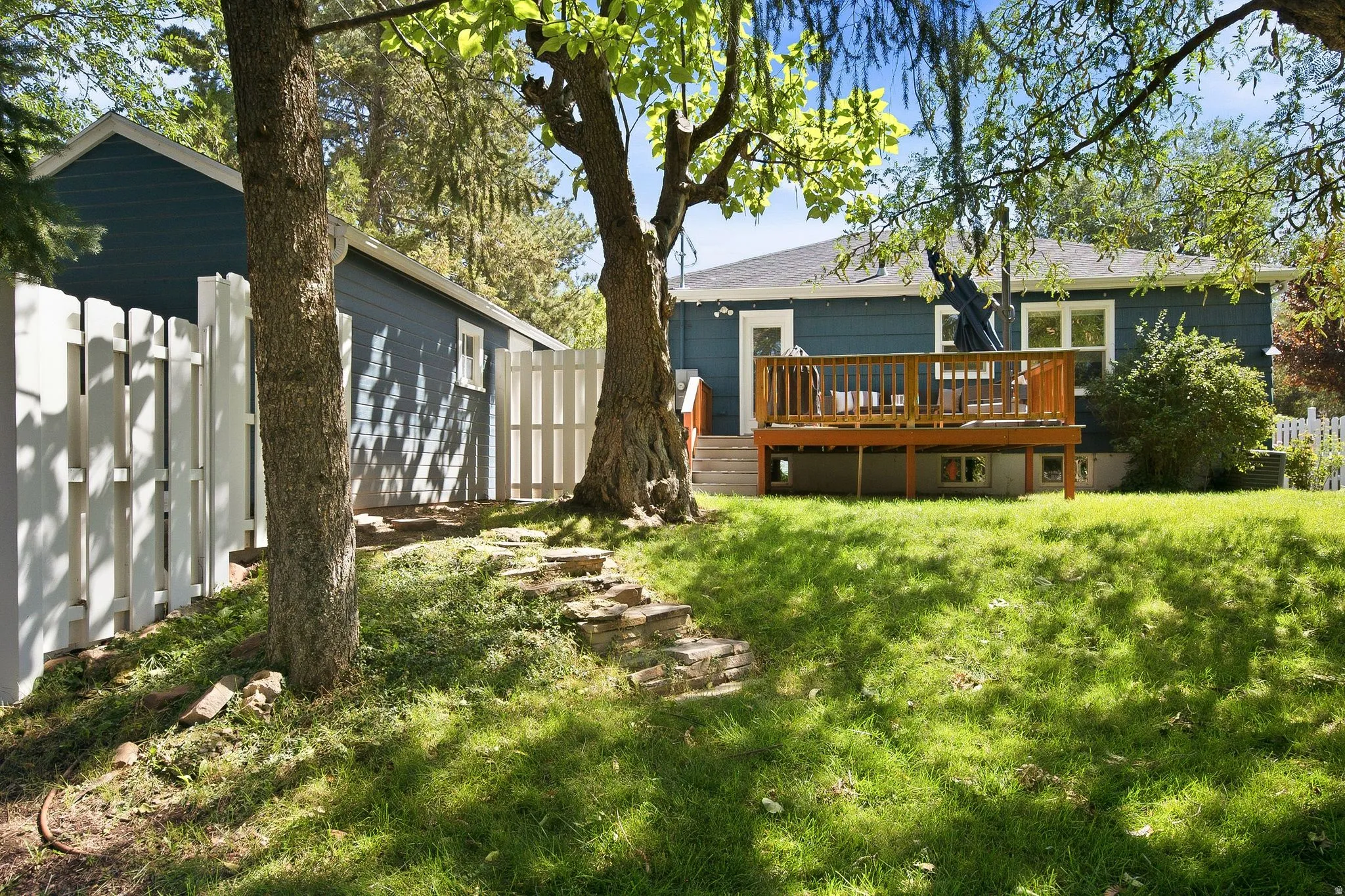 Mature shade trees anchor this expansive backyard oasis, featuring a manicured lawn, white picket fencing, and a wood-deck addition with railings