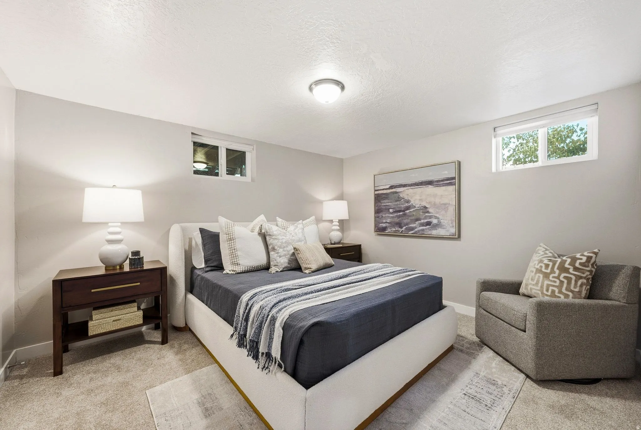 Spacious bedroom suite featuring a platform bed with upholstered headboard, wood-finish nightstands with integrated lighting, neutral carpeting, recessed ceiling fixture, and an upholstered accent chair