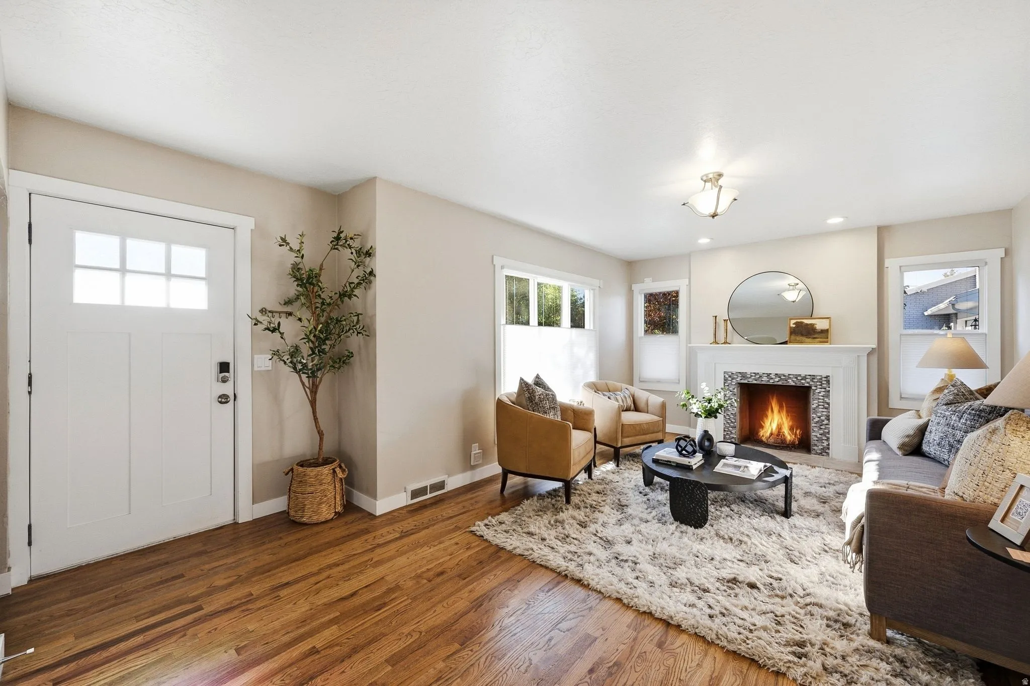 Welcoming entry foyer with white paneled door and sidelights opening to a spacious living area featuring hardwood flooring, a fireplace with surround mantel, recessed ceiling lighting, and large windows allowing natural illumination throughout the open-co