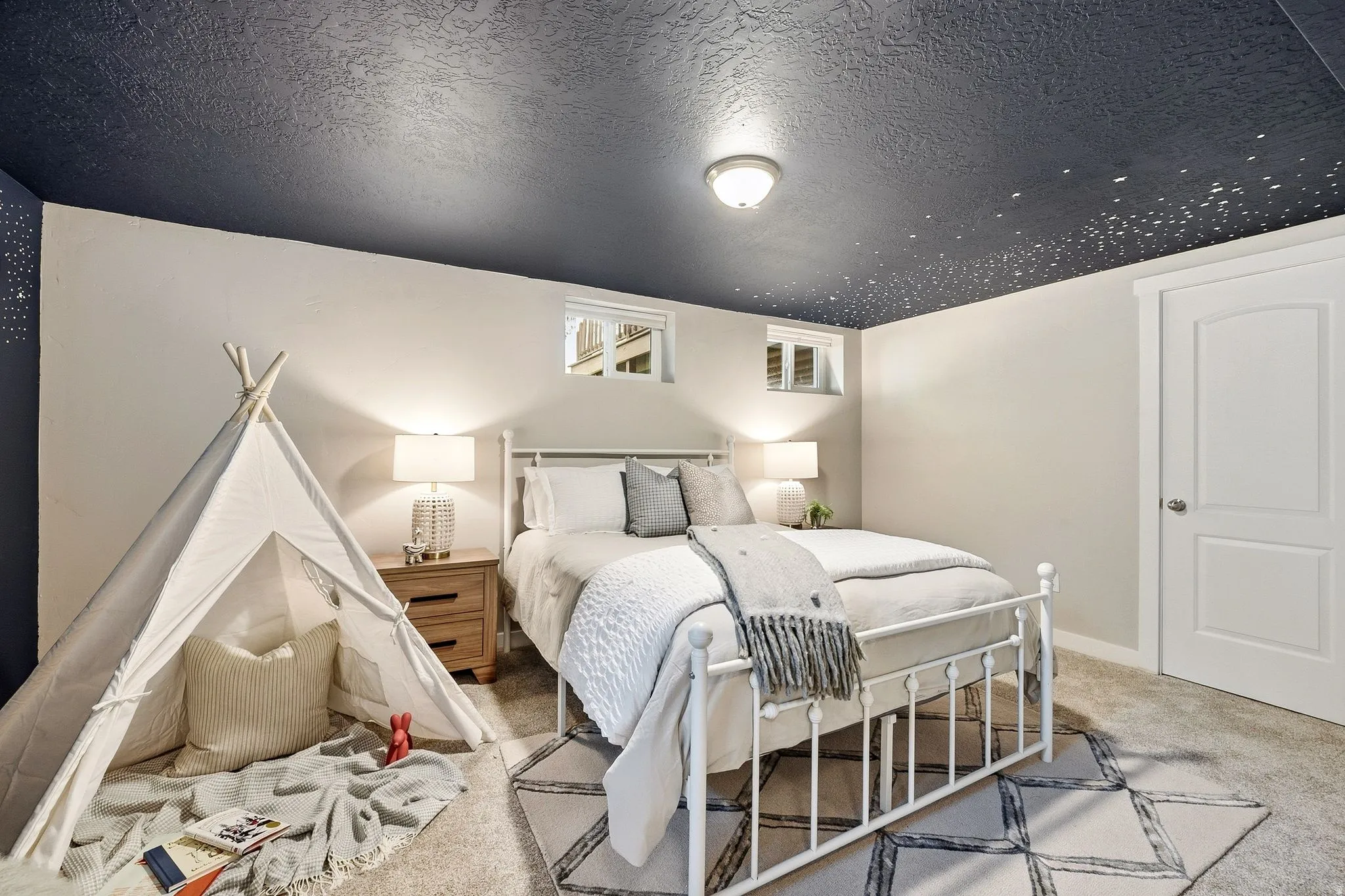 Bright, light-filled bedroom featuring a metal bed frame, dual bedside tables with attached lighting, wood-finish flooring, and geometric area rug