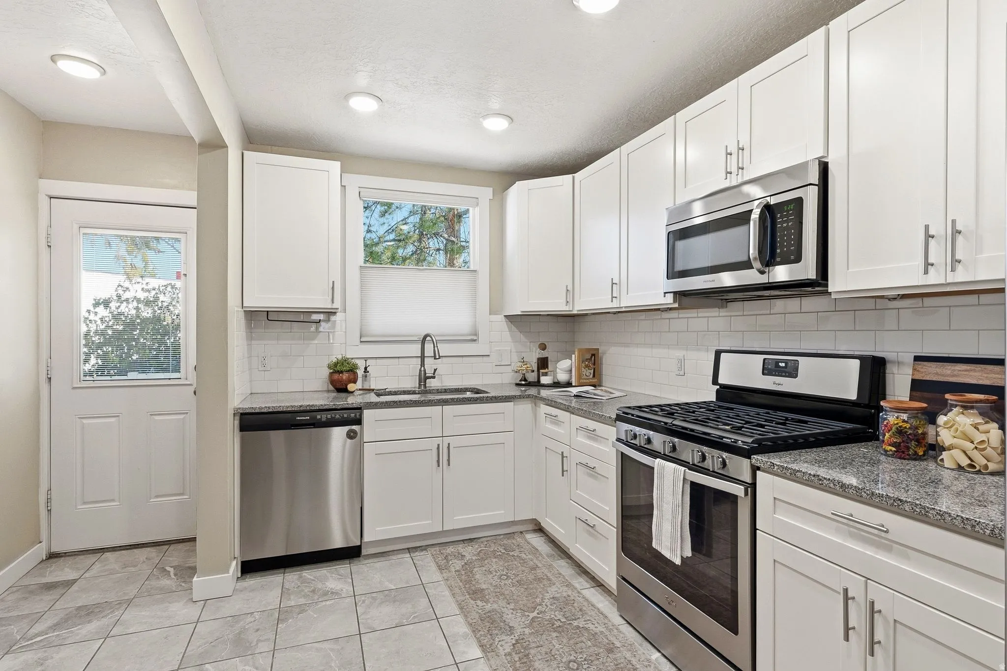 Bright, updated kitchen featuring white shaker cabinetry, stainless steel appliances including a gas range and built-in microwave, granite countertops, subway tile backsplash, and tile flooring