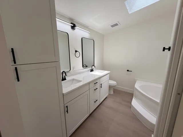 Dual vanity with white cabinetry and integrated sinks, complemented by a skylight, corner soaking tub, and wood-finish flooring