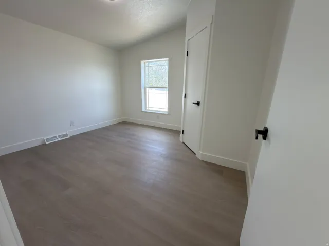 Light-filled room featuring wood-finish flooring, white walls, a white door with matte black hardware, and a window with blinds