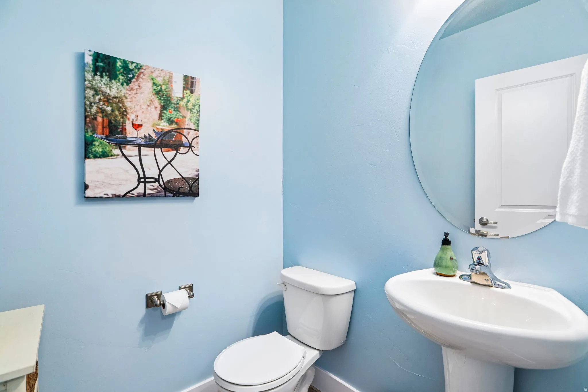 Bright, light-filled powder room featuring soft blue walls, a pedestal sink with single-handle faucet, a low-profile toilet, and a large round mirror