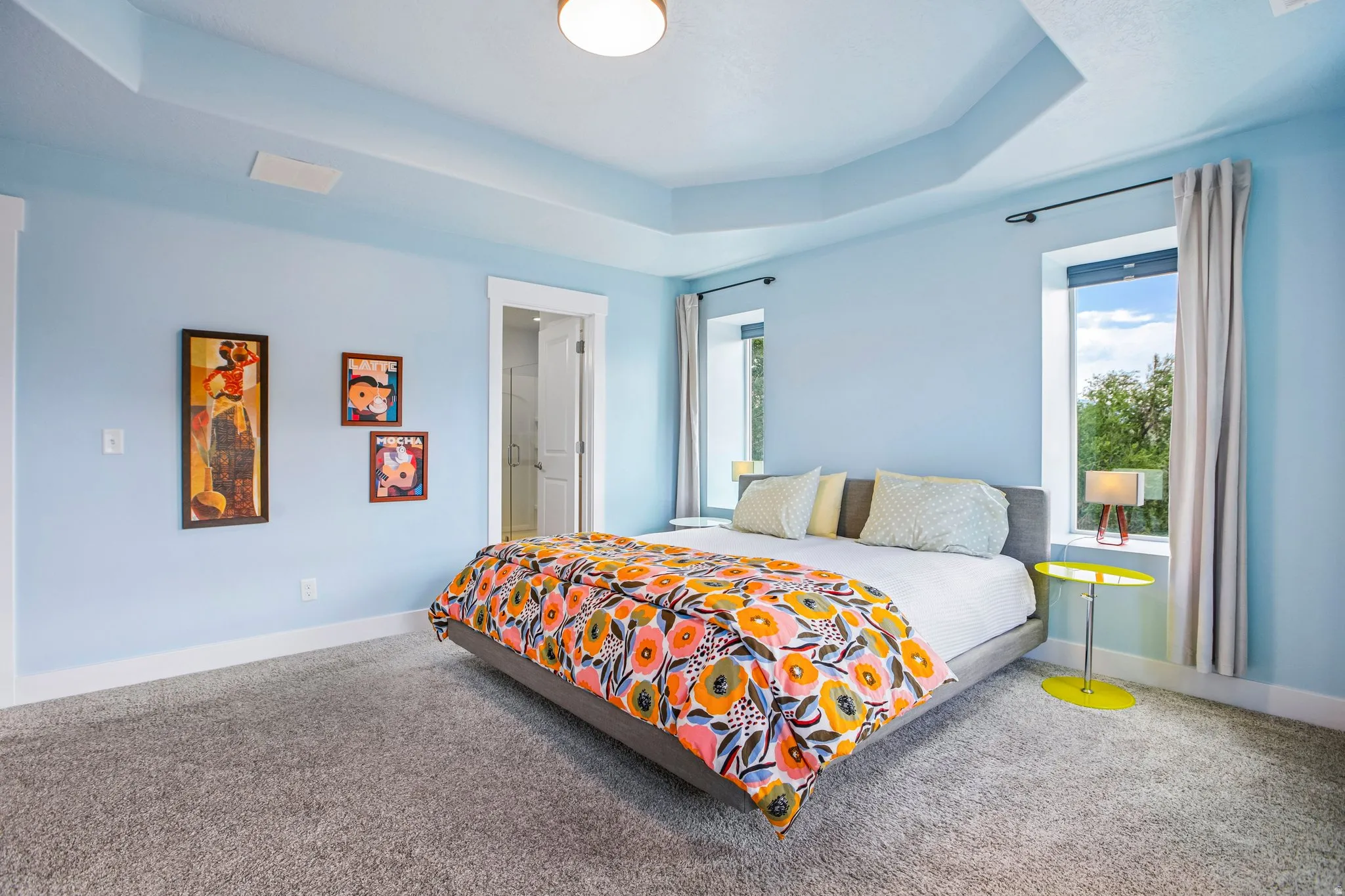 Spacious primary bedroom showcasing a coffered ceiling with recessed lighting, soft blue walls, gray carpeting, and expansive windows with natural light