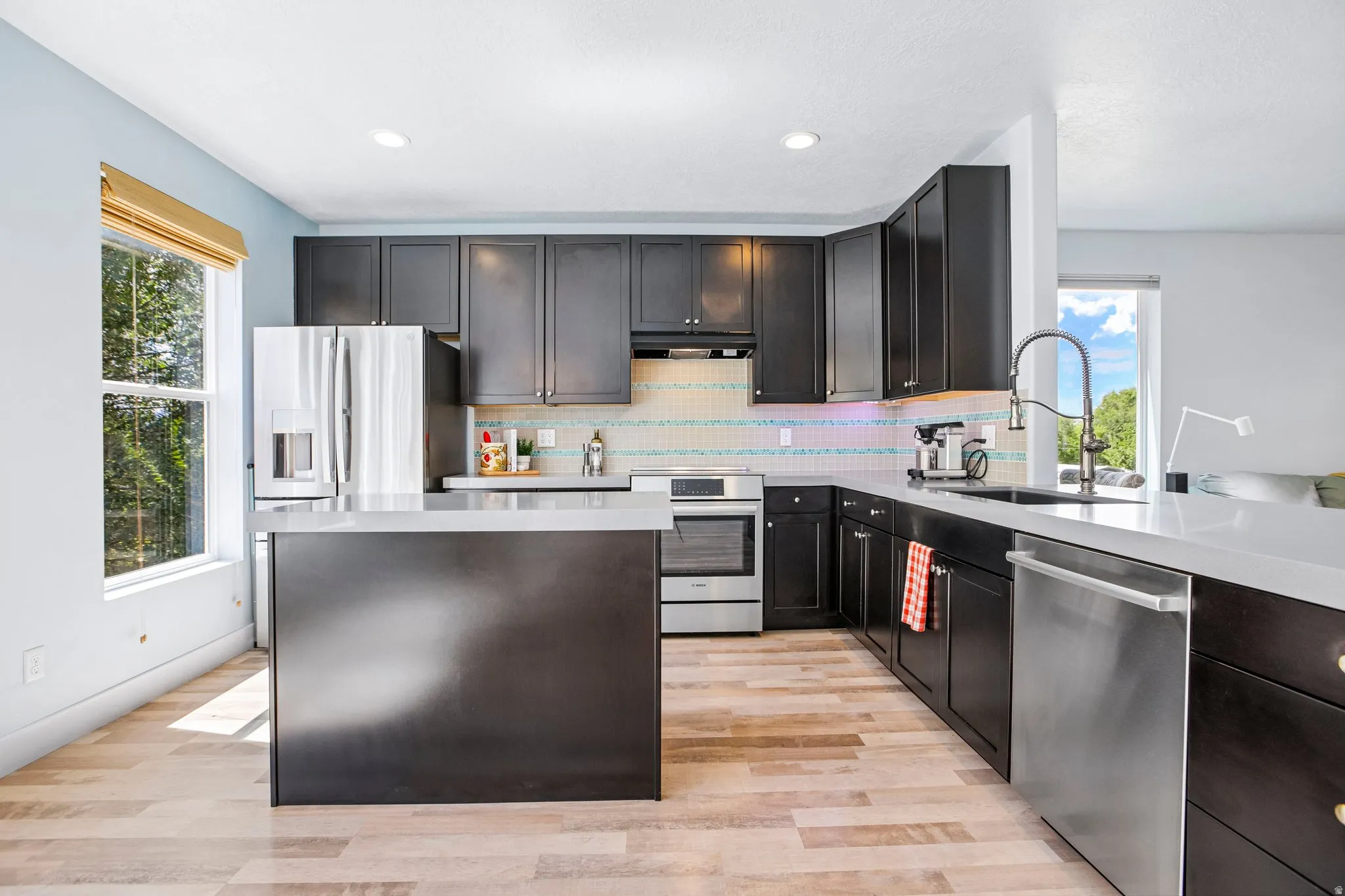 Modern kitchen featuring dark cabinetry with stainless steel appliances, a large waterfall-edge island with seating, white subway tile backsplash, quartz countertops, and wood-finish flooring throughout the open layout